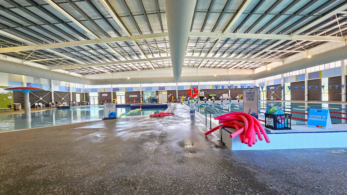 To the left is a leisure pool with a water feature structure. Next to it is the spa pool. On the right is an 8-lane, 25m swimming pool. The floor is non-slip concrete, and bench chairs are placed on the left. Red hose reel,, banners, and safety reminders, including blue triangle banners, are displayed around the pool. The high ceiling has insulation foams and features large air-conditioning vents, metal beams, and bright lights.