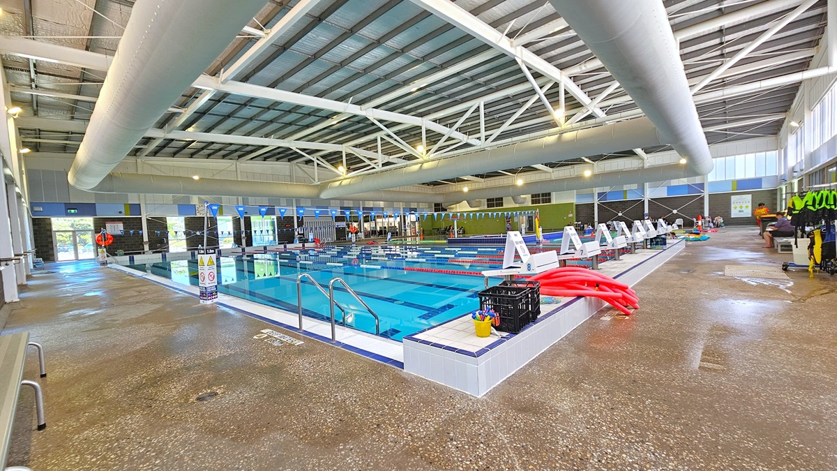 The 8-lane, 25m swimming pool features handrails and eight diving blocks. Swim accessories like red noodle hoses are on the pool deck. On the left, there is bench seating, and on the right, a rack for hanging swimming gear. The floor is non-slip concrete, and safety reminders, including blue triangle banners, are displayed. The high ceiling has insulation foams and large air-conditioning vents, metal beams, and bright lights.