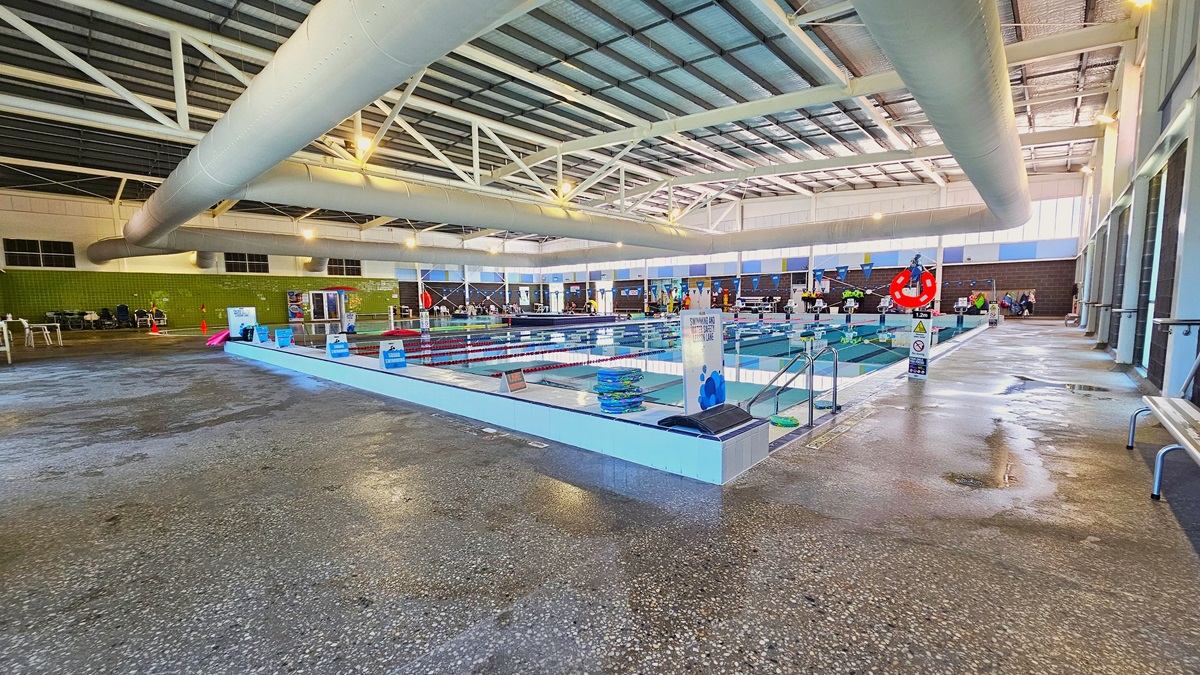 The 8-lane, 25m swimming pool has handrails in the corner, with swim accessories like red noodle hoses and floaters on the pool deck. On the right, there is bench seating, and on the left, there are aquatic wheelchairs aligned against the green tiled wall. The floor is non-slip concrete, and safety reminders, including blue triangle banners, are displayed. The high ceiling features insulation foams, large air-conditioning vents, metal beams, and bright lights.