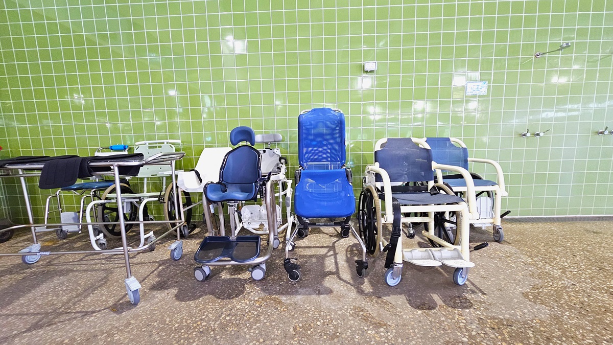 Aquatic wheelchairs and a cart with towels are positioned against the green-tiled wall, adjacent to the showers. The floor is made of non-slip concrete.