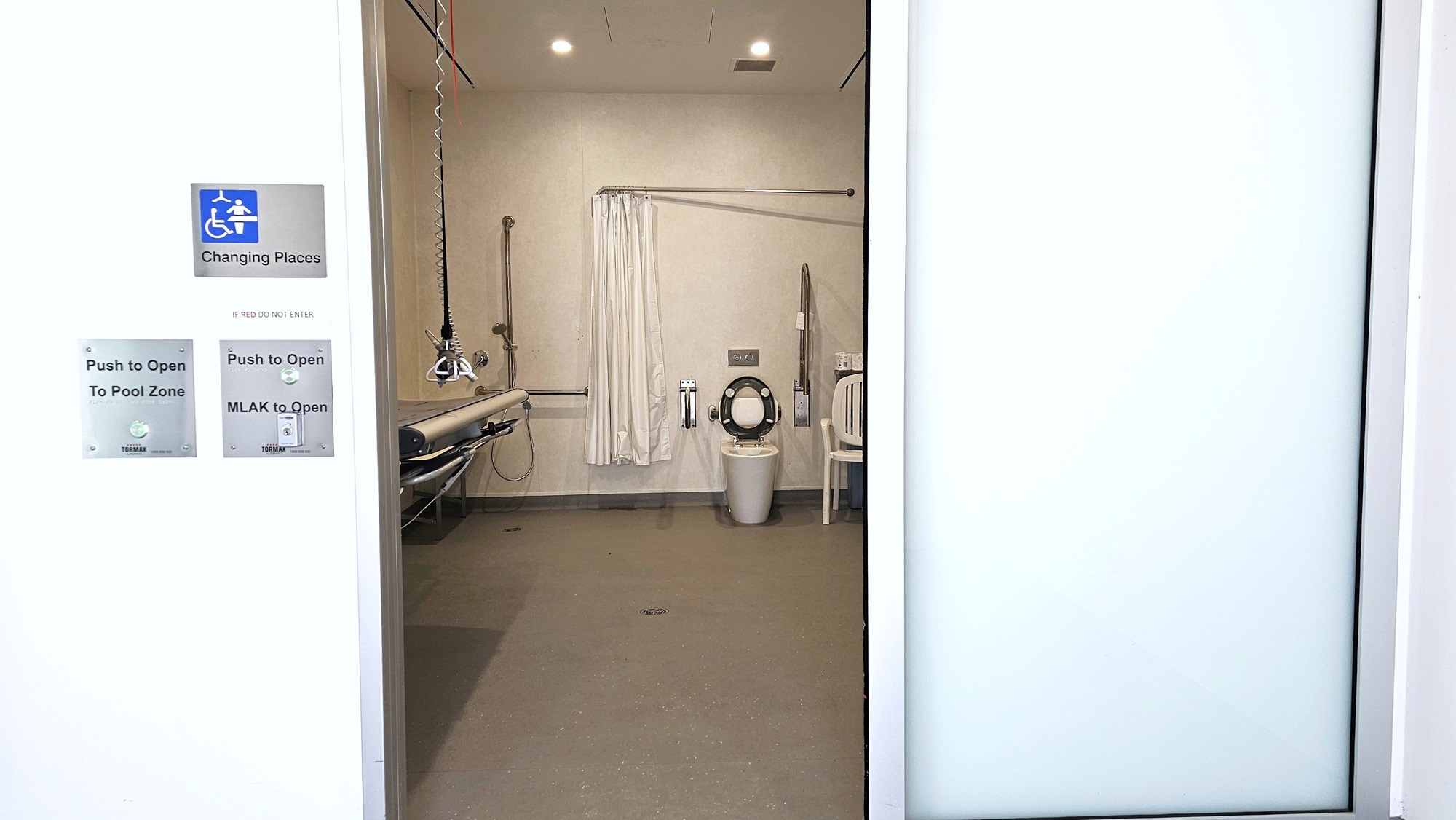 The Changing Places bathroom is accessible with an MLAK key, indicated on the door sign. An adult-size changing table with a hoist is situated on the left, and on the right, there is a toilet bowl with handrails and a nearby plastic chair. To the left are showers with a white curtain.