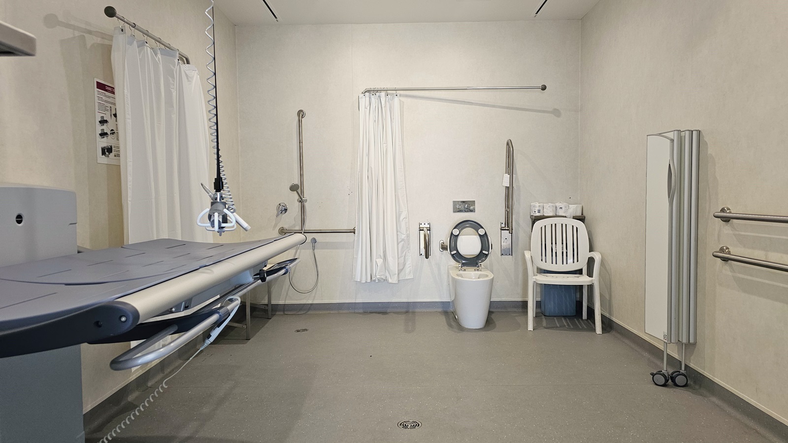 Inside the Changing Places, an adult-size changing table with a hoist is on the left, along with a shower area featuring white curtains and handrails. In the middle is a toilet bowl with handrails, accompanied by a white plastic chair and nearby tissue rolls. On the right, there is a collapsible partition, additional handrails affixed to the wall, and a white-themed interior with a grey matte floor.