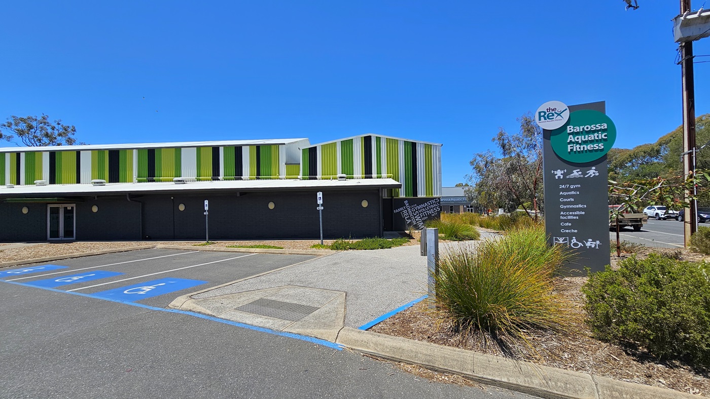 The parking area is situated near the curb, featuring five accessible parking spaces. A sign displaying the venue's name is located on the concrete plant box adorned with trees and ornamental plants. The building showcases an industrial design with alternating hues of green, yellow, white, and black. A tall lamp post stands beside an electrical post. Several cars are parked along the street.