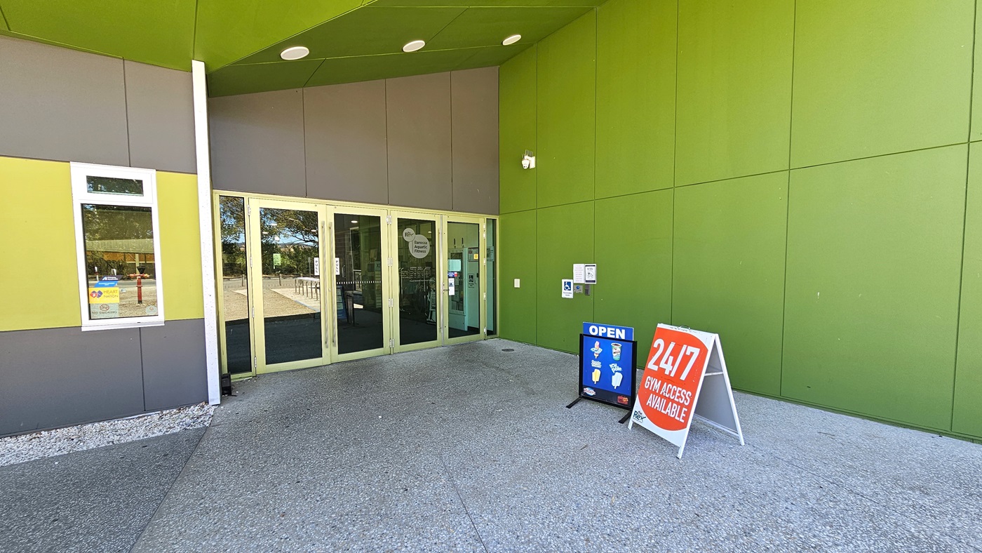 Access to the facility is through manually operated, outward-opening glass doors with cream trim. The ceiling features an elevating design with three circular fluorescent lights. The wall on the right is painted green, including the ceiling. To the left is a window. Freestanding banners bearing notices and reminders are visible on the right.