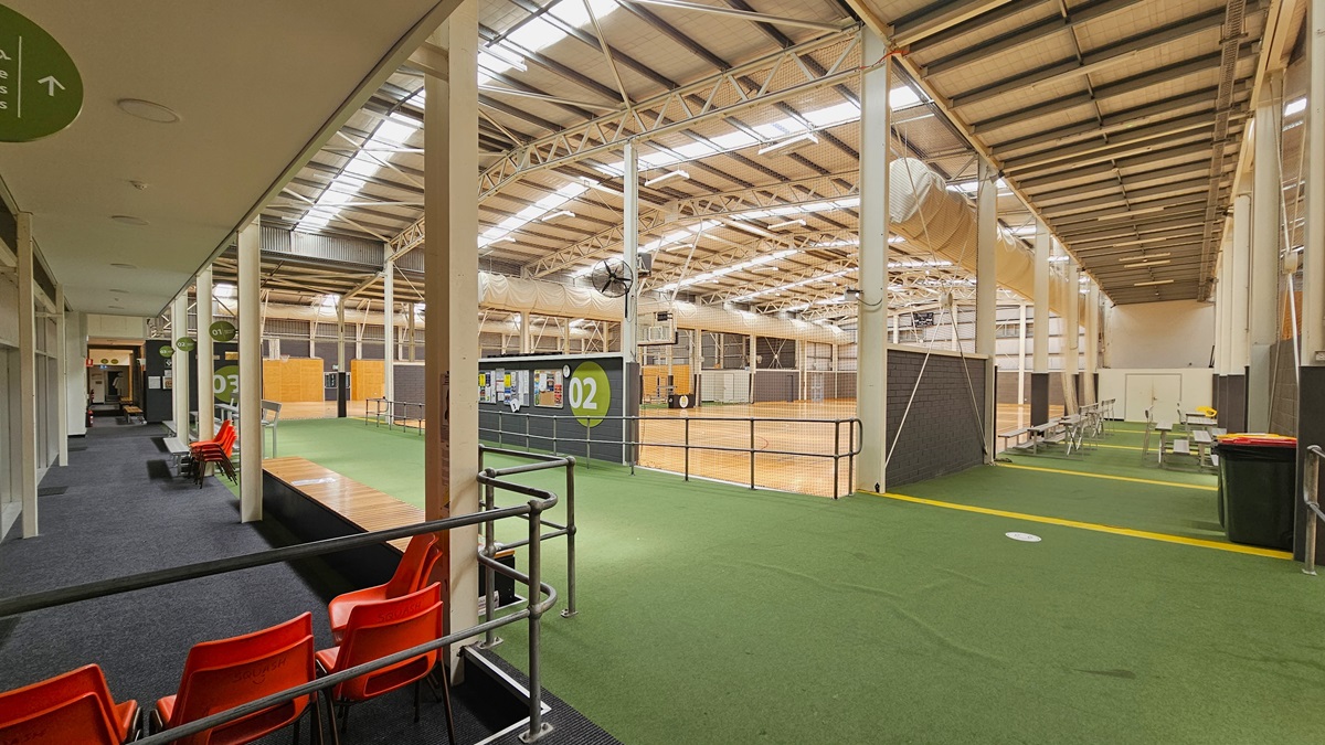 The basketball court features a carpeted area on the left with tiered and bench seating, enclosed by metal rails. In the center, a spacious basketball court with timber floors and netted walls is present. On the right, another carpeted area offers plastic movable chairs for seating. The section displays a wooden panel ceiling.