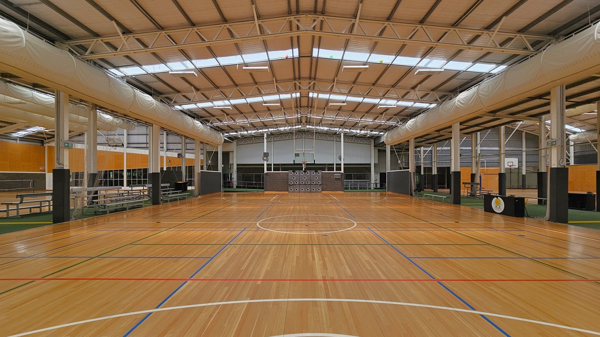 The court has a spacious area with timber floors marked for ball games. The ceiling has an industrial look with exposed beams, large air conditioning vents, fluorescent lights, and natural skylights. On the left, there is an area with green carpets and tiered seating. On the right, another area with green carpets is equipped with bench chairs and tables. The surrounding wall has a wooden design.