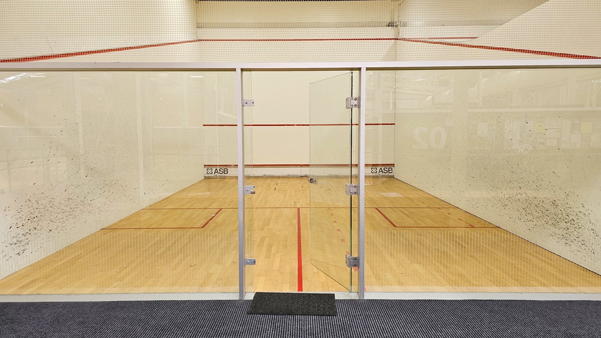 The squash court has a timber floor with red markings, enclosed by a net and stainless steel barriers. White walls with black markings surround the court, and there is a glass door in the middle that opens inward, serving as the entry point. The middle wall features an inscription "ASB" on the lower section.