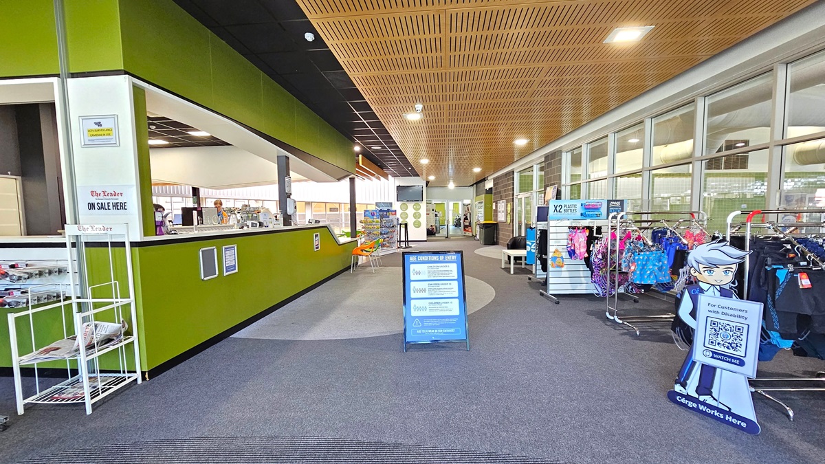The facility's entrance is through a manually operated outward-opening door with cream trim. A sticker bearing the facility's name is on the glass wall. The right wall is green, and the upper wall is light grey. Inside, a section of the retail and reception area is visible.