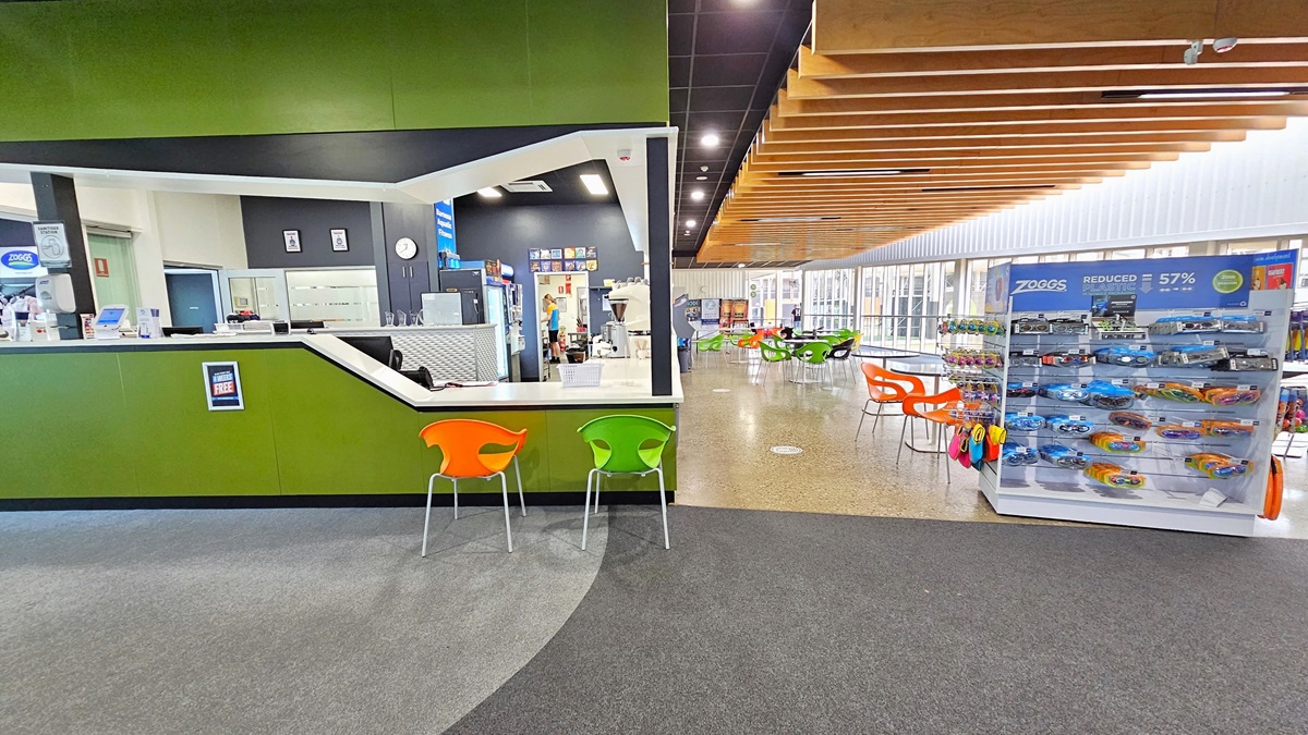 To the left is the reception with green walls and a brown ceiling. The reception counter has a sloping design for seated assistance. An alcohol dispenser and a card-swiping machine are placed on the counter. Orange and yellow-green plastic chairs are positioned for seating. A display rack with goggles and swimming accessories is on the right. Movable plastic tables are placed in front of the cafe. The floor is tiled with granite, and the ceiling has a timber wooden panel design.