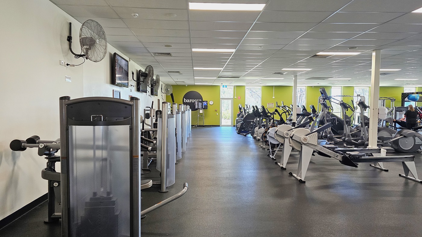 Access to the gymnasium is through a manual glass door that opens inward. Reminders are affixed to the glass door. Two large freestanding banners advertising the facility are positioned on both sides. Inside, a section of the gym is visible, including display racks on the left and training machines.