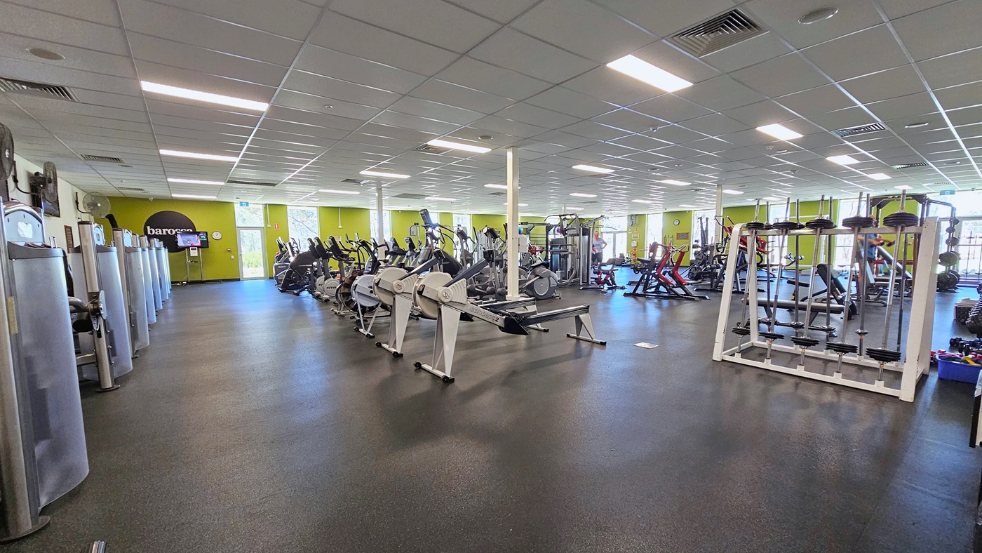Upon entering the gym, exercise machines line the left wall, accompanied by flat screen TVs and a wall fan. In the middle, additional machines are positioned along with weightlifting machines. The floor is covered in rubber tiles, and the ceiling features a white porous design with centralized air conditioning. At the far end, a green wall is complemented by floor-to-ceiling windows. Rubber mats cover the floor, and the ceiling, adorned with a porous design, is illuminated by fluorescent lights.