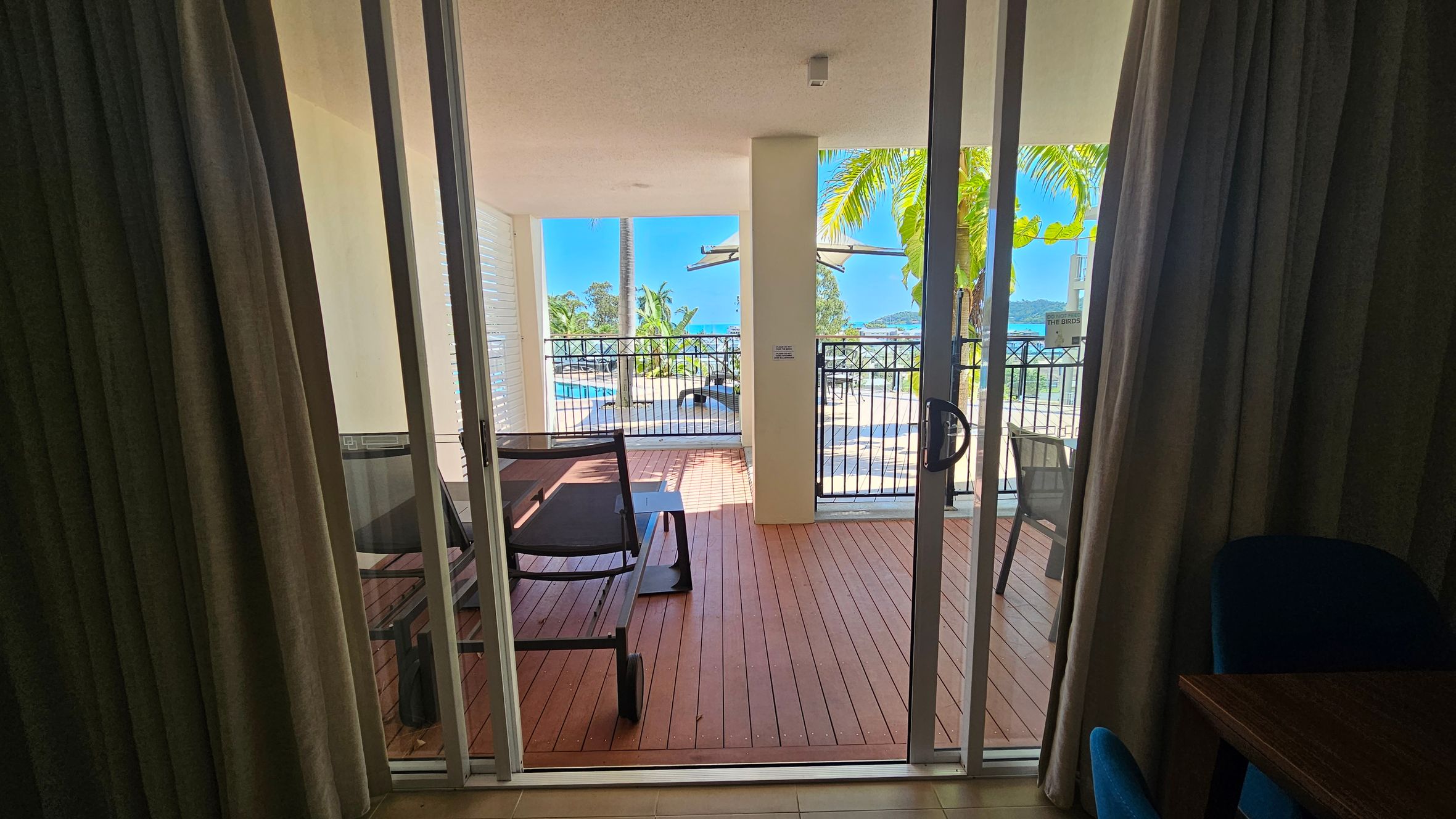 A view through glass sliding doors leading to a wooden deck. On the deck, there are two lounge chairs and a small table. Beyond the deck, a black metal railing encloses the area. In the background, palm trees and a swimming pool are visible, with a glimpse of the ocean and distant hills. Curtains frame the doors on either side, and part of a wooden table with a blue chair is visible on the right.