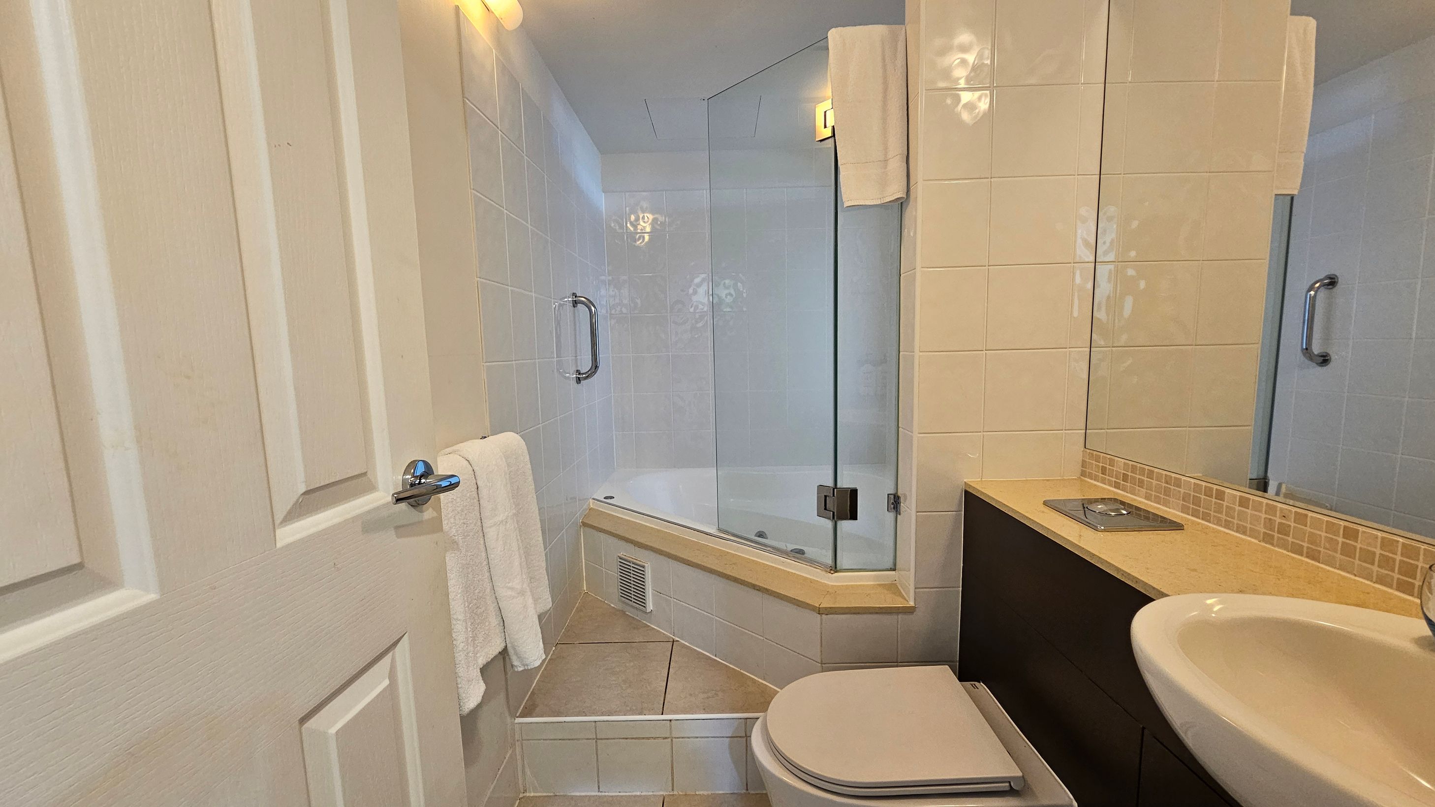 A small bathroom with a white door on the left, featuring a towel hanging on a chrome handle. The centre has a glass-enclosed bathtub with a chrome grab bar and a towel draped over the glass. On the right, there's a beige countertop with a built-in sink and a large mirror above. Below the counter is a dark cabinet. The floor is tiled in a light colour, and the walls are covered with white tiles.