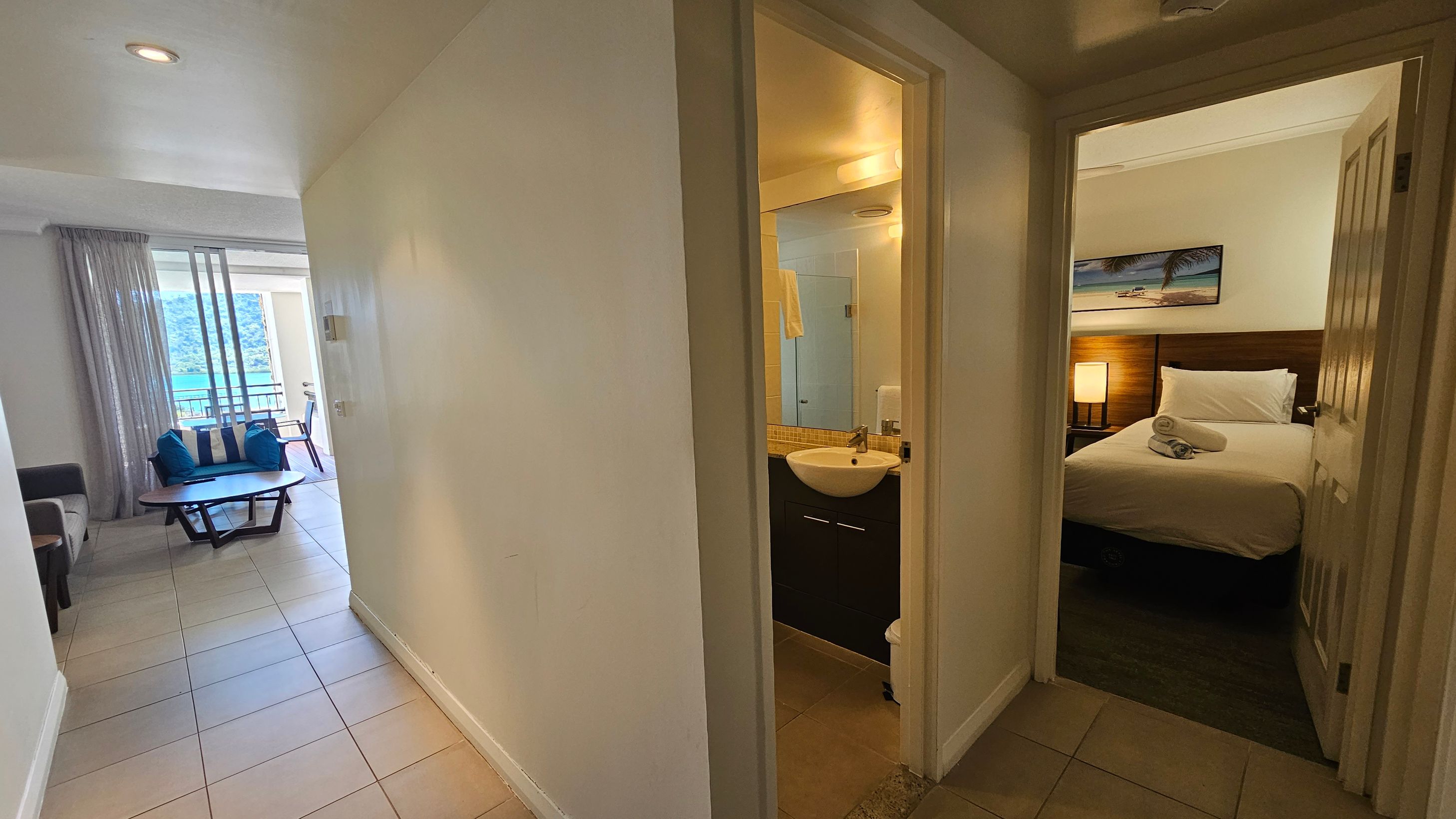 A hallway with tiled flooring leads to a living area on the left, featuring a sofa, a coffee table, and a view of a balcony with outdoor chairs. Sheer curtains cover the windows. On the right, a bathroom with a sink and mirror is visible. Further right, a bedroom with a neatly made bed, a bedside lamp, and a beach-themed picture on the wall. The ceiling is plain with recessed lighting.