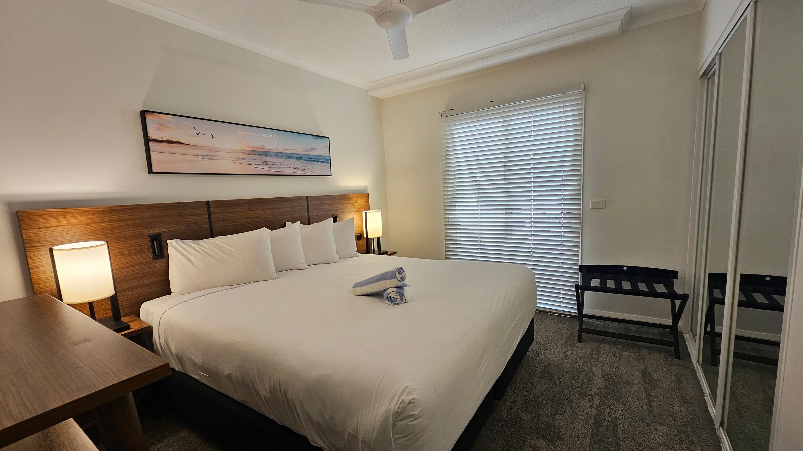 A bedroom with a large bed in the centre, covered with a white duvet and two rolled towels on top. The headboard is wooden, flanked by two bedside tables with lamps. Above the bed is a landscape painting. On the right, a window with closed blinds and a luggage rack beneath. A mirrored wardrobe is also on the right. The floor is carpeted in green, and a ceiling fan is visible above.