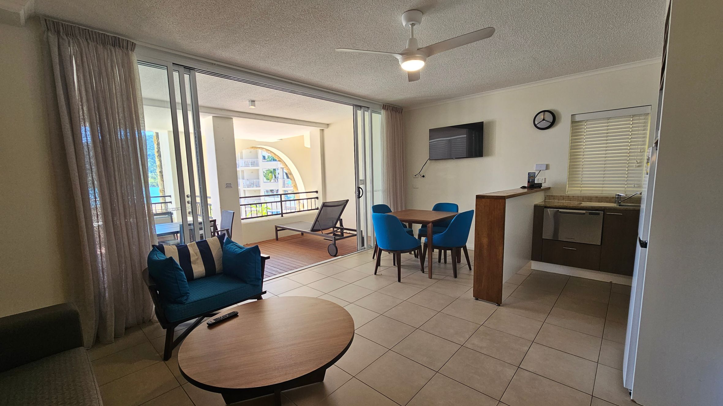 Living area with tiled flooring and a ceiling fan. On the left, a blue armchair with striped cushions sits beside a round wooden coffee table. Sliding glass doors lead to a balcony with a lounge chair and outdoor table. On the right, a small dining table with four blue chairs is near a wall-mounted TV. A kitchen area with a sink, oven, and fridge is adjacent, with a window featuring blinds and a wall clock above.