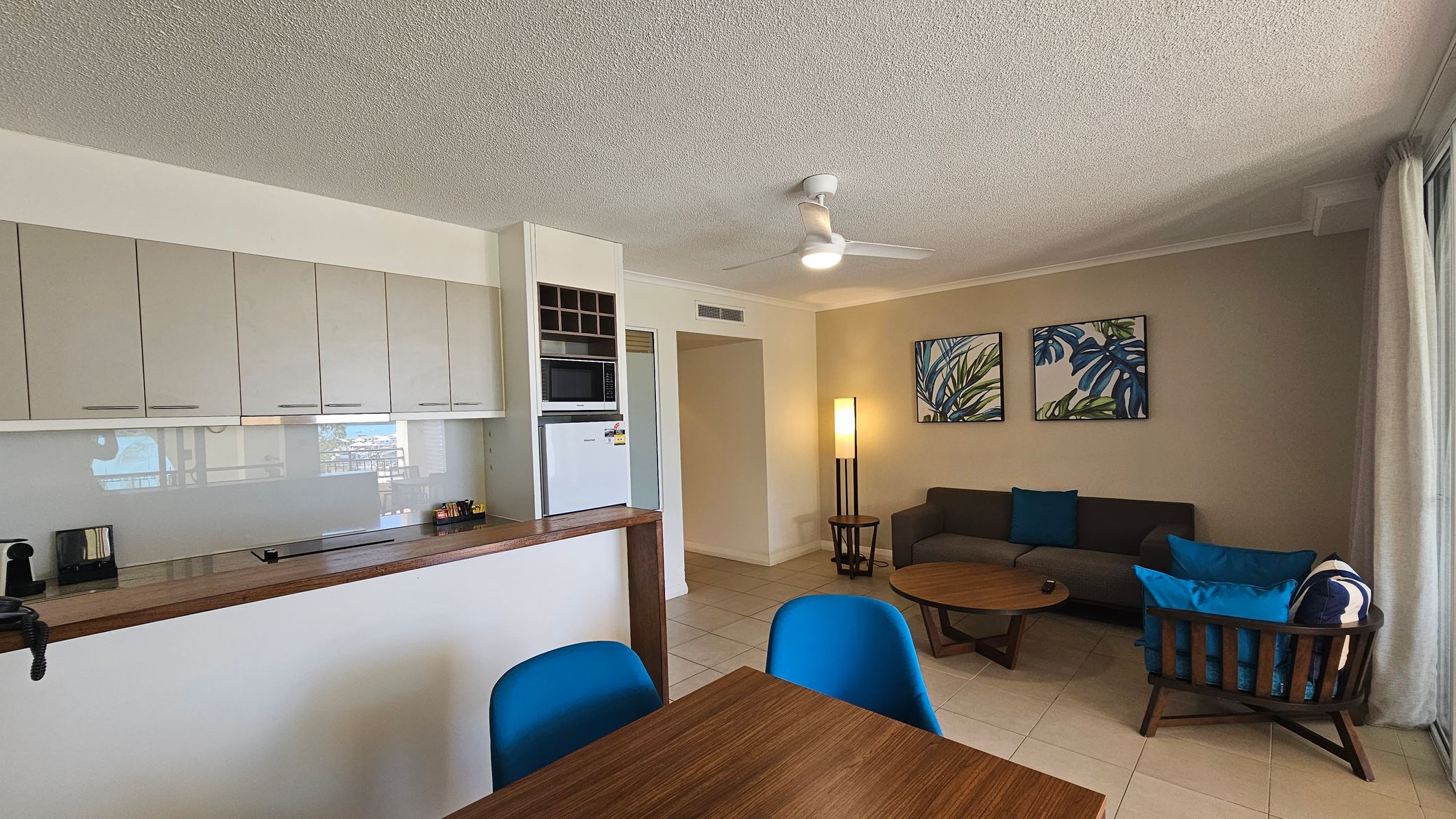 A modern living space with a kitchen on the left featuring beige cabinets, a microwave, and a fridge. A wooden counter holds a toaster and other small items. In the centre, a wooden dining table with blue chairs. On the right, a seating area with a grey sofa, blue cushions, and a wooden coffee table. Two botanical prints hang on the wall above the sofa. A tall floor lamp stands nearby. The room has tiled flooring and a ceiling fan. Large windows with curtains are on the far right.