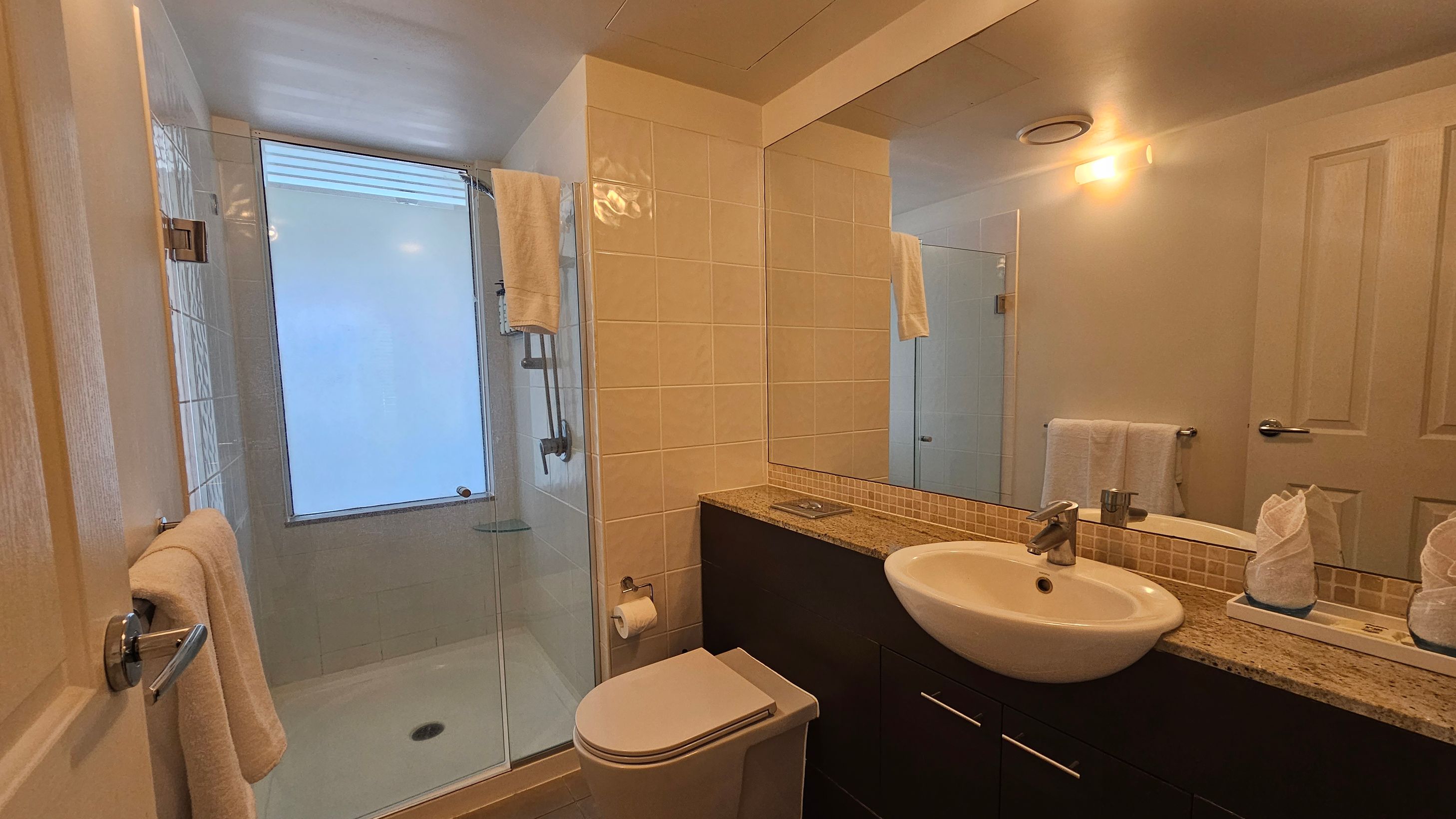 A bathroom with a glass-enclosed shower on the left, featuring a frosted window and a towel hanging on the wall. The centre has a toilet with a roll of toilet paper mounted on the wall. On the right, a granite countertop with a white sink and a large mirror above. Two neatly folded towels are placed on the counter. The walls are tiled, and the ceiling has a recessed light fixture. A door with a towel hanging on it is partially visible.