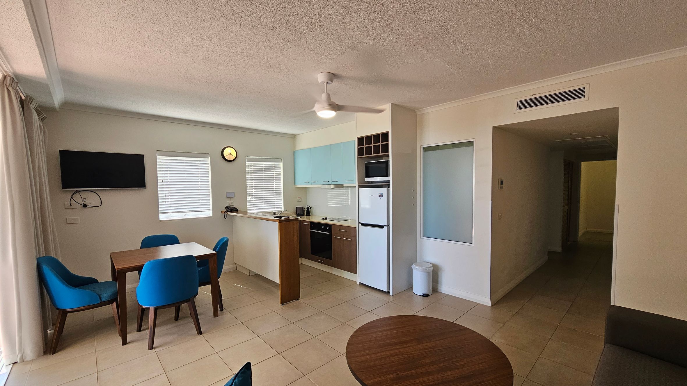 A modern living space with a tiled floor. On the left, there's a dining table with four blue chairs and a wall-mounted TV above. The kitchen area is in the middle, featuring light blue cabinets, an oven, a microwave, and a fridge. A small clock is on the wall above the kitchen counter. On the right, there's a hallway leading to another room. A ceiling fan is centrally located, and a round wooden table is partially visible in the foreground.