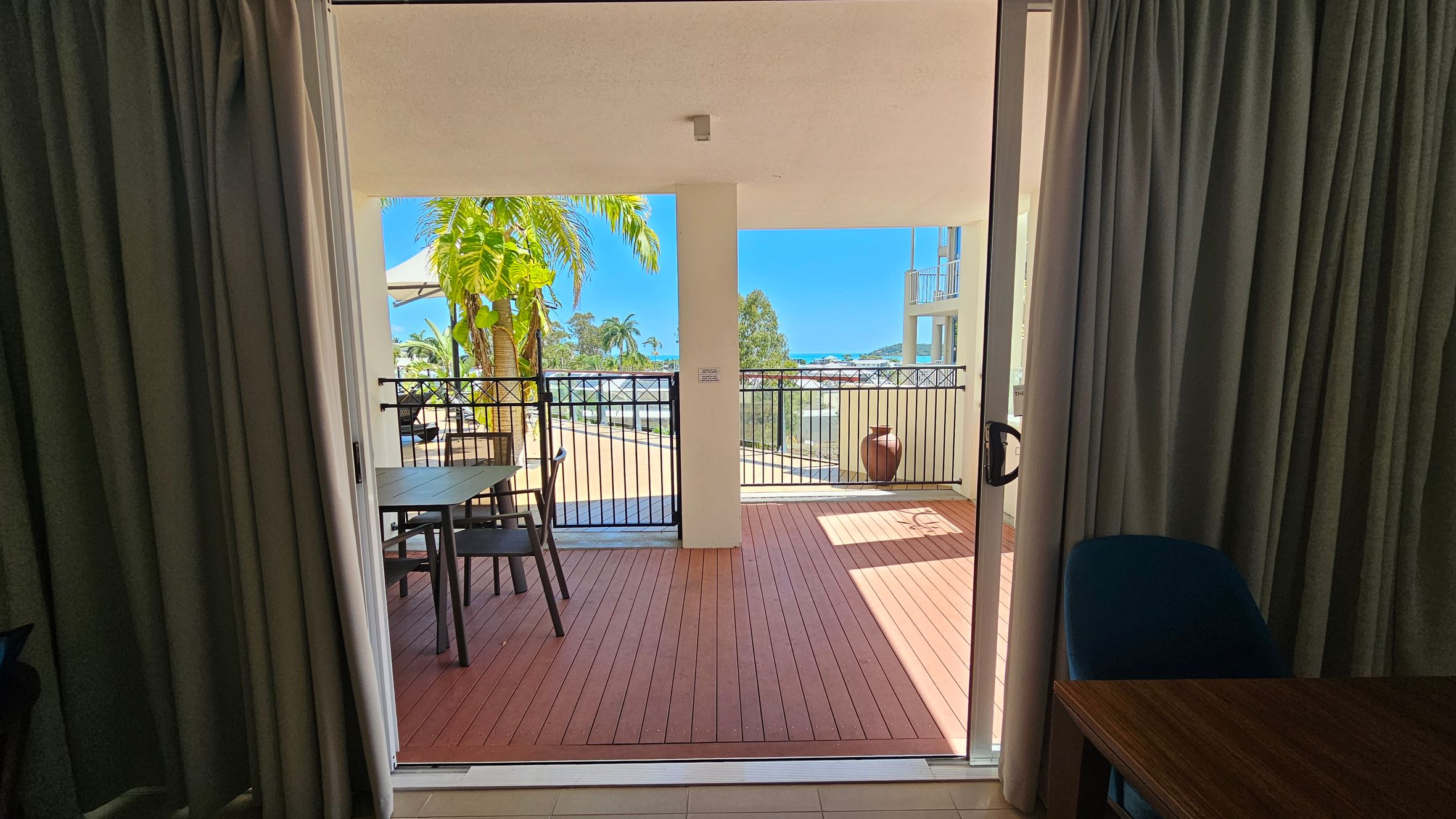 View from inside a room looking out onto a balcony. The foreground has dark green curtains on both sides and a wooden table with a blue chair on the right. The balcony features a wooden floor, a black metal railing, and a large potted plant on the right. A table with chairs is on the left side of the balcony. In the background, there are palm trees and a glimpse of the sea and distant buildings under a clear blue sky.