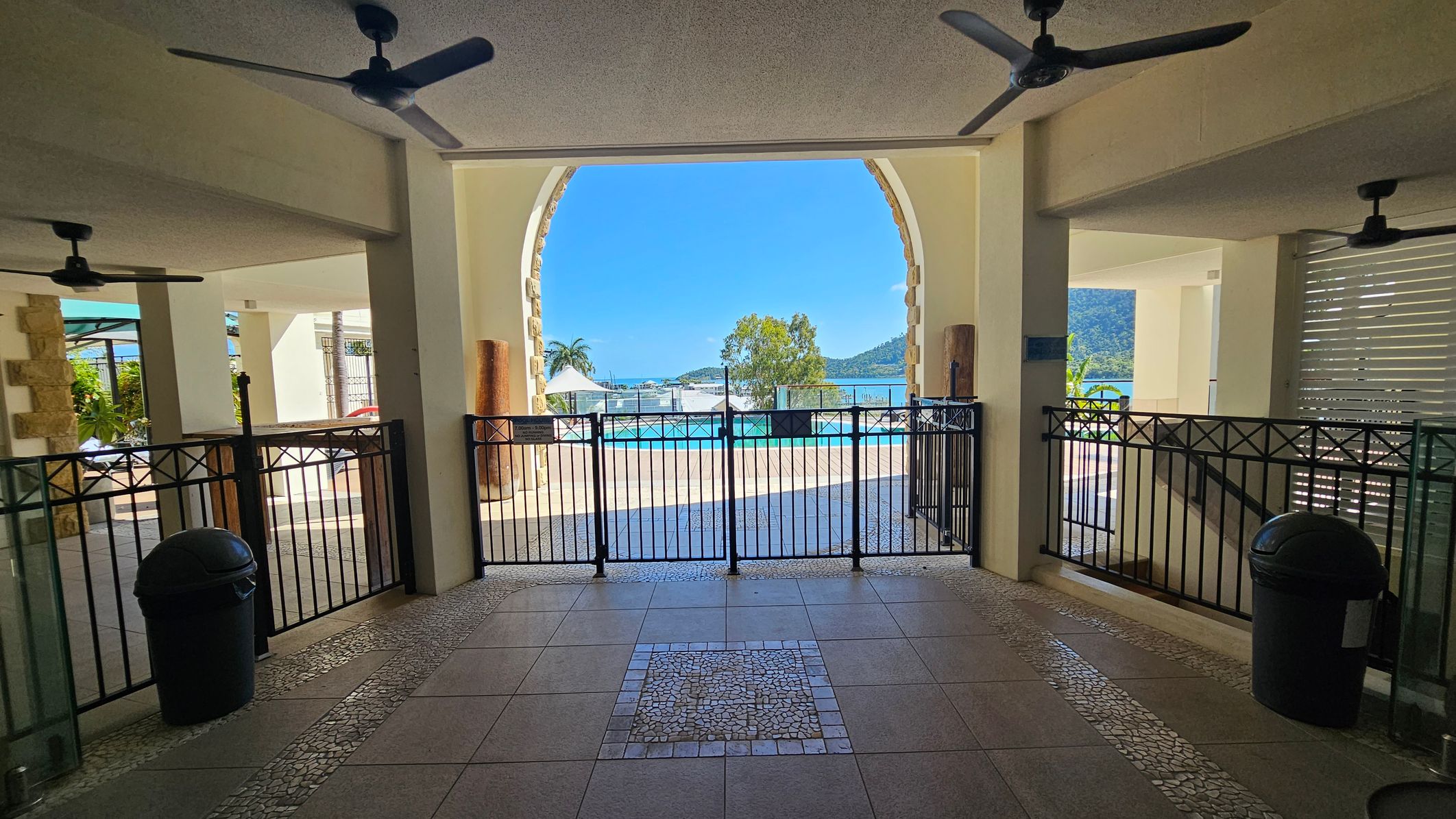 A view through an archway with a black metal gate leading to an outdoor pool area. The foreground has a tiled floor with a mosaic pattern. Ceiling fans are visible above. On the left and right, there are black bins and stone pillars. Beyond the gate, a pool is visible with a backdrop of trees, a palm tree, and distant hills under a clear blue sky.