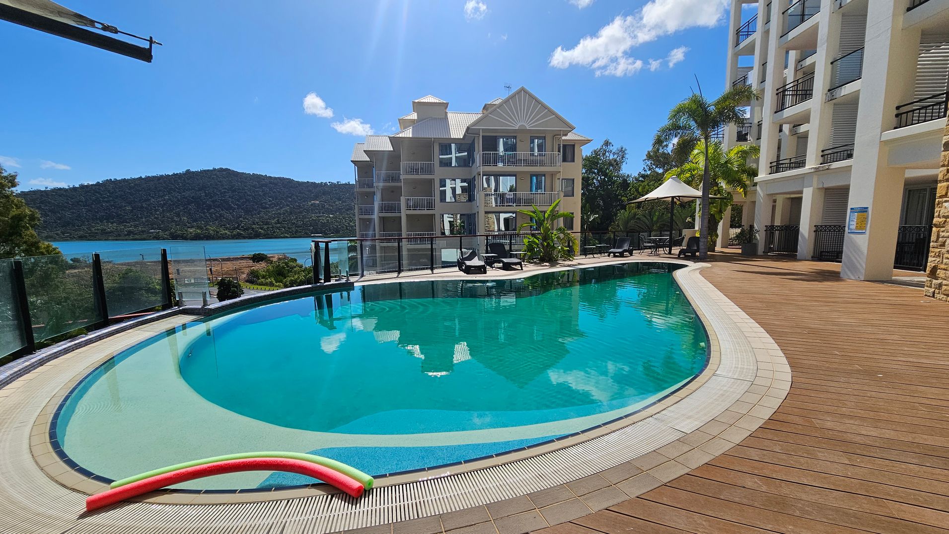 A curved swimming pool with clear blue water is surrounded by a wooden deck. On the right, there are several sun loungers and a white umbrella. A multi-storey building with balconies is visible in the background. To the left, a glass fence overlooks a body of water and a tree-covered hill. A red and yellow pool noodle rests on the pool's edge. Palm trees and other greenery are scattered around the area.