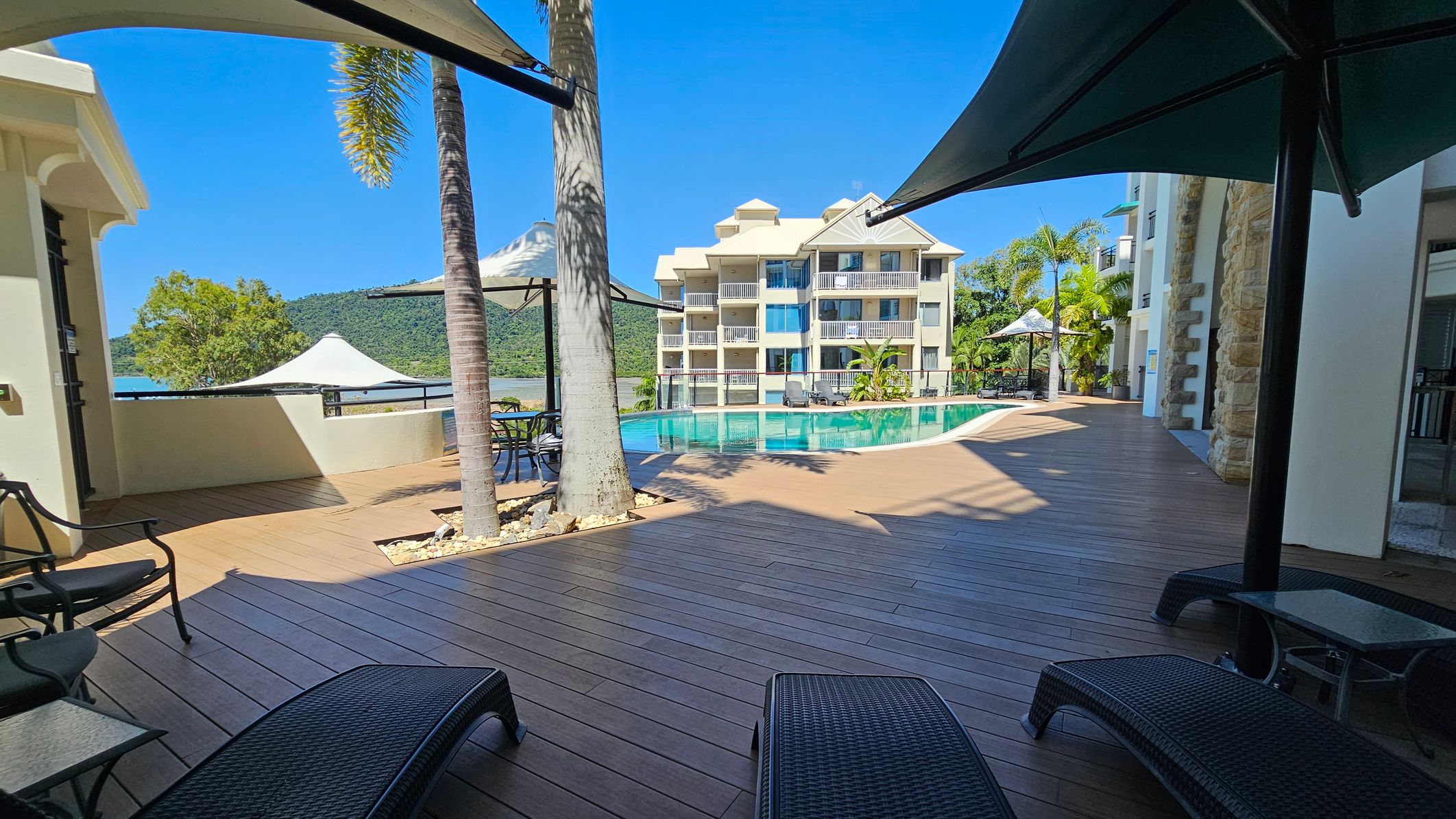 A poolside area with wooden decking and several sun loungers in the foreground. On the left, there are chairs and a table under a large umbrella. A tall palm tree stands nearby. In the centre, a clear swimming pool reflects the surrounding buildings. In the background, a multi-storey building with balconies is visible, along with lush green hills and a glimpse of water. On the right, another umbrella provides shade over more seating.