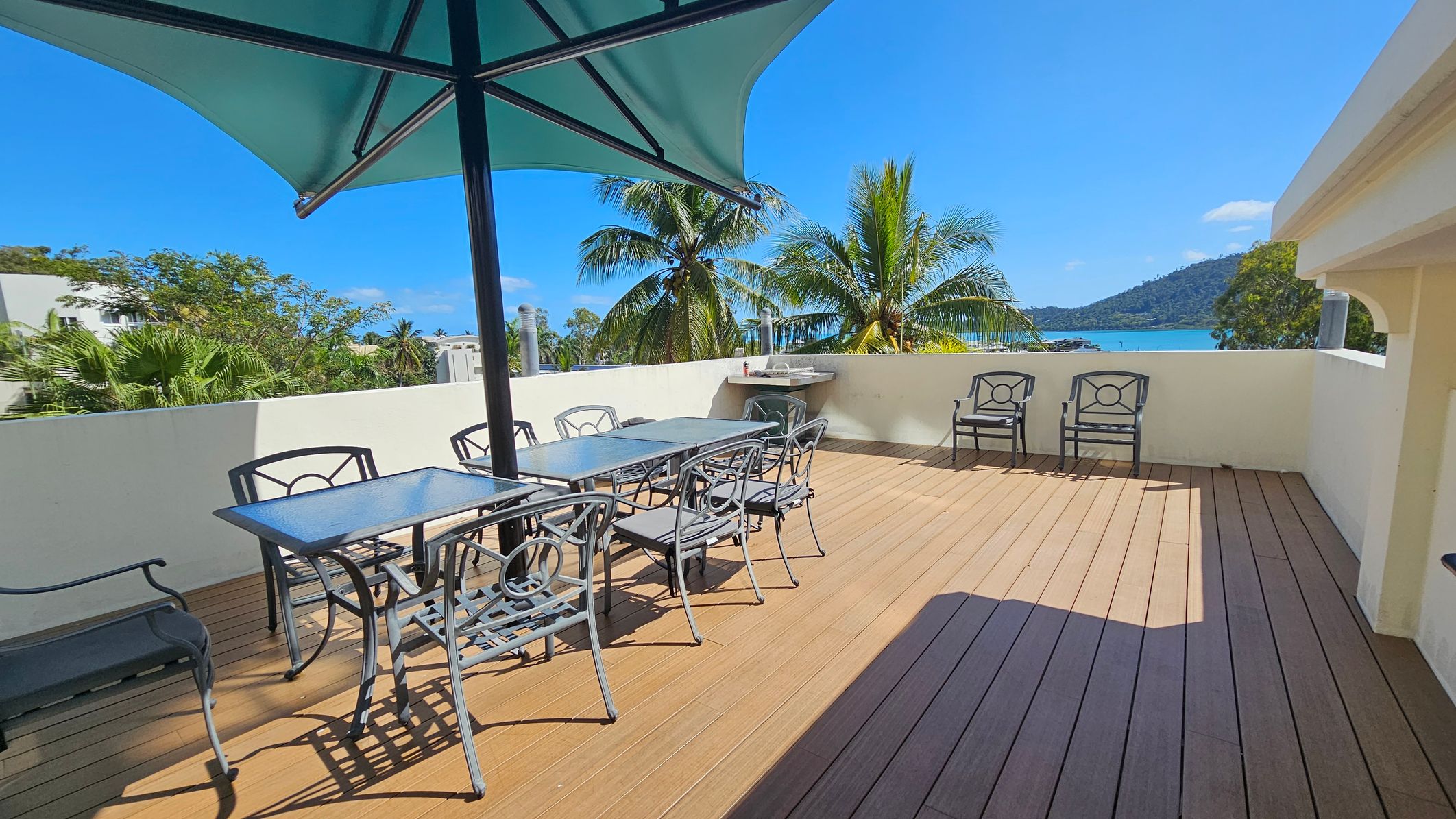 A spacious outdoor terrace with wooden flooring. On the left, there's a large table with six metal chairs under a green umbrella. In the background, palm trees are visible, and beyond them, a view of the sea and a distant hill. On the right, two additional chairs are placed against a low white wall. A small outdoor sink or counter is also visible near the wall.
