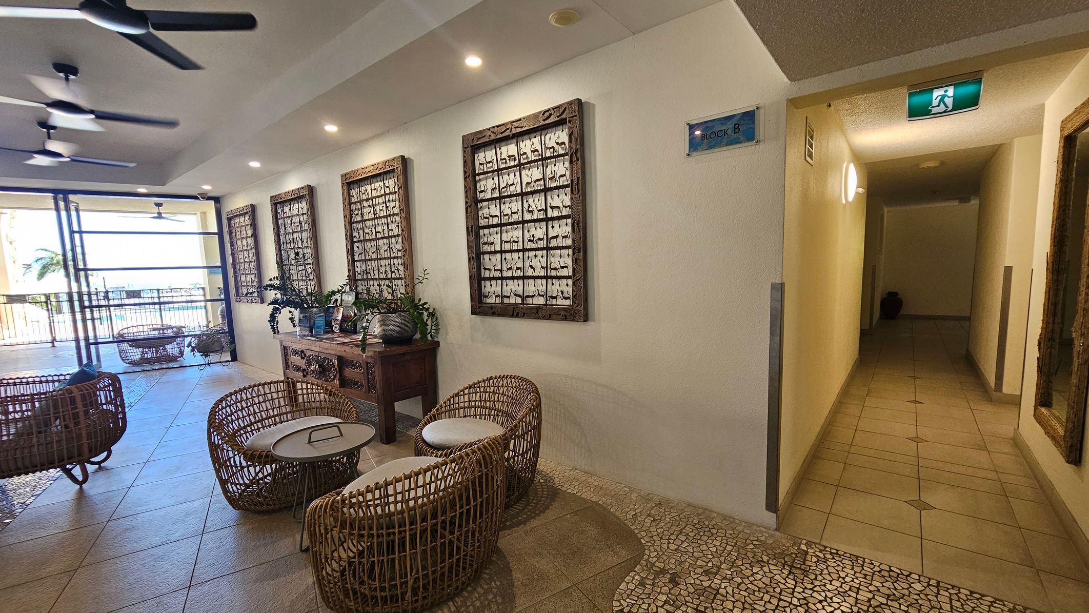 A spacious indoor area with wicker chairs and small tables on a tiled floor. On the left, large windows open to a balcony with a view of palm trees and the sea. The ceiling has multiple ceiling fans. The right wall features three framed artworks and a wooden console table with plants. A hallway with a tiled floor and an illuminated exit sign is visible on the far right.