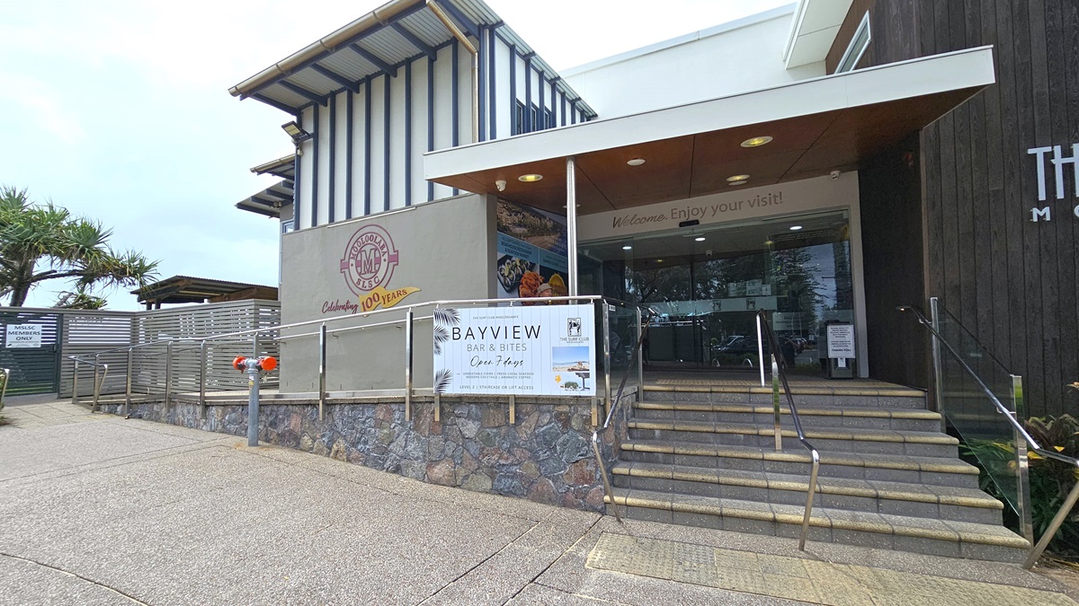 A wide-angle view of the entrance to a modern building labelled "Mooloolaba." A flight of stairs with handrails and truncated domes leads up to glass double doors, flanked by stone walls. A ramp with railings is on the left. A wall-mounted sign reads 'Welcome, enjoy your visit!' Below it, a banner displays 'BAYVIEW Bar & Bites'. Palm trees are visible on the left.