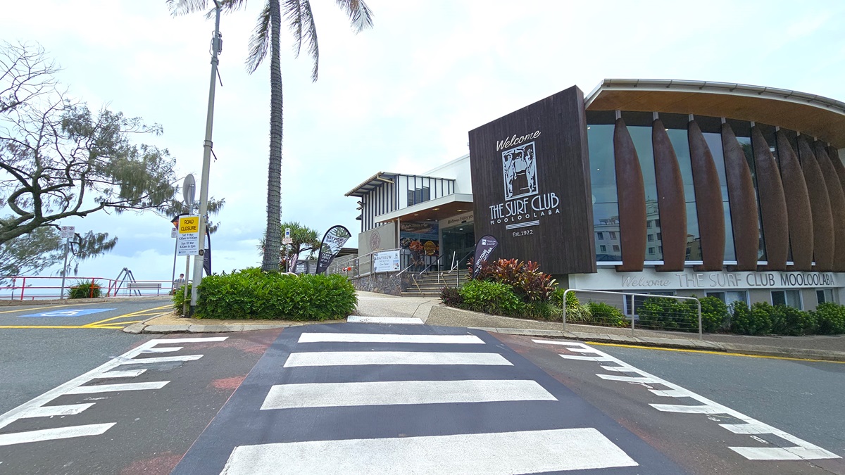 In the foreground, a pedestrian crossing leads to a building with a prominent sign reading "The Surf Club Mooloolaba". The architecture showcases dark tones with vertical wood panelling. Entry to the building is provided by stairs and a ramp with handrails. Palm trees and landscape plants flank the front and sides of the building. The ocean fills the backdrop.