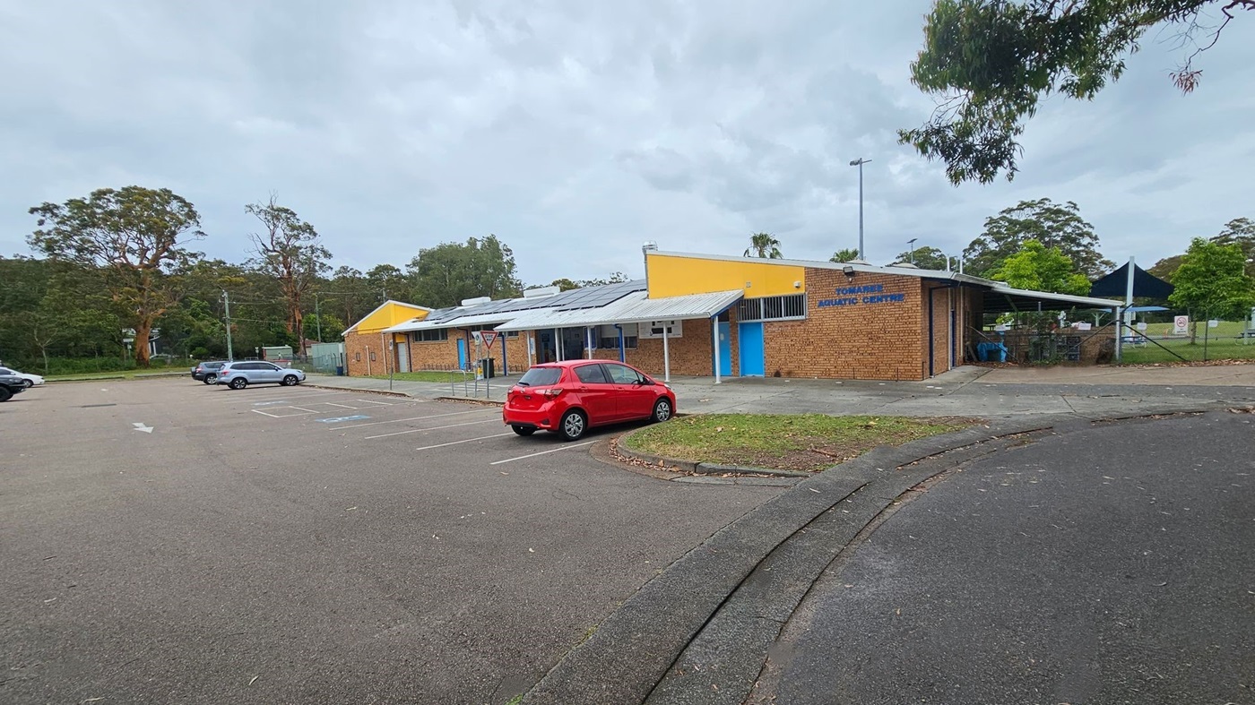A brick building with a yellow and blue facade is on the right, labelled "Aquatic Centre". A red car is parked in front of it. To the left, there are several parking spaces with a few cars. The background features tall trees and greenery. The foreground shows a paved area with some scattered leaves.