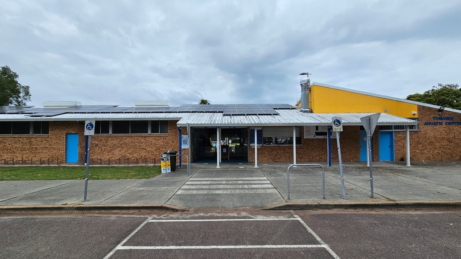 A brick building with a metal roof featuring solar panels. The entrance is in the centre, flanked by blue doors on either side. A yellow section of the building is on the right. There are signs for disabled parking in front, with a zebra crossing leading to the entrance. The foreground has a paved area and parking spaces, with some fallen leaves scattered around. Trees are visible in the background.