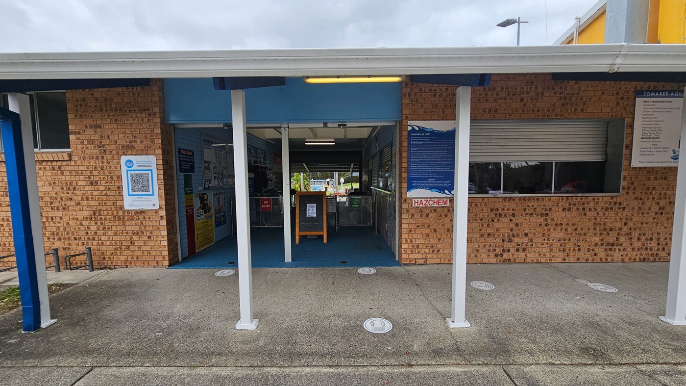 A brick building with a covered entrance supported by white pillars. On the left, a QR code sign is displayed on the wall. The entrance is open, revealing a blue interior with various signs and a wooden stand. To the right, a "HAZCHEM" sign and a shuttered window are visible. The ground is a mix of green and grey concrete, and there are circular markers on the floor. A sign with information is mounted on the right wall.
