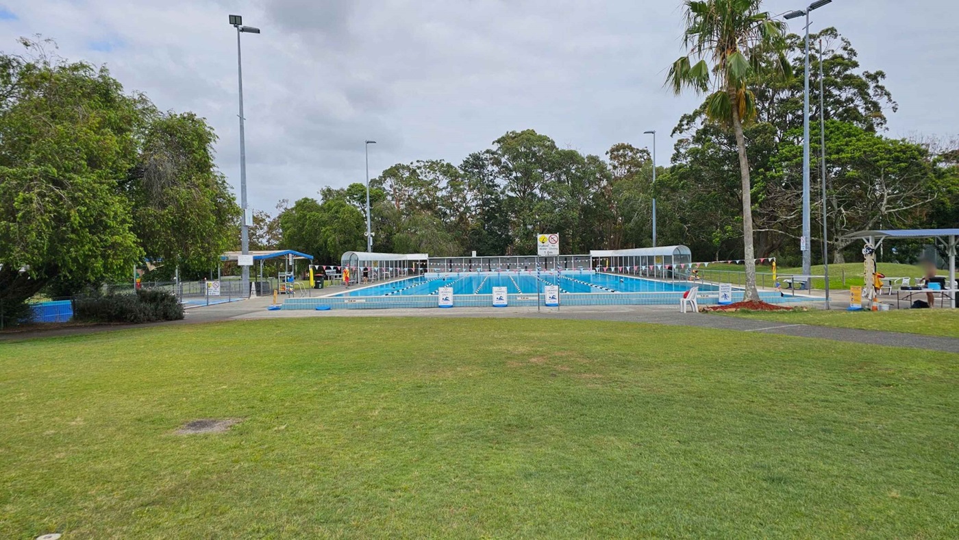 A park with a concrete path leading towards a swimming pool area. On the left, there is a large blue water slide structure surrounded by trees. In the centre, two tall palm trees stand near the path. On the right, a pool is covered by dark triangular shade sails, with signs and benches nearby. The grass is green and well-maintained, and there are landscaped flower beds near the path. Trees line the background.