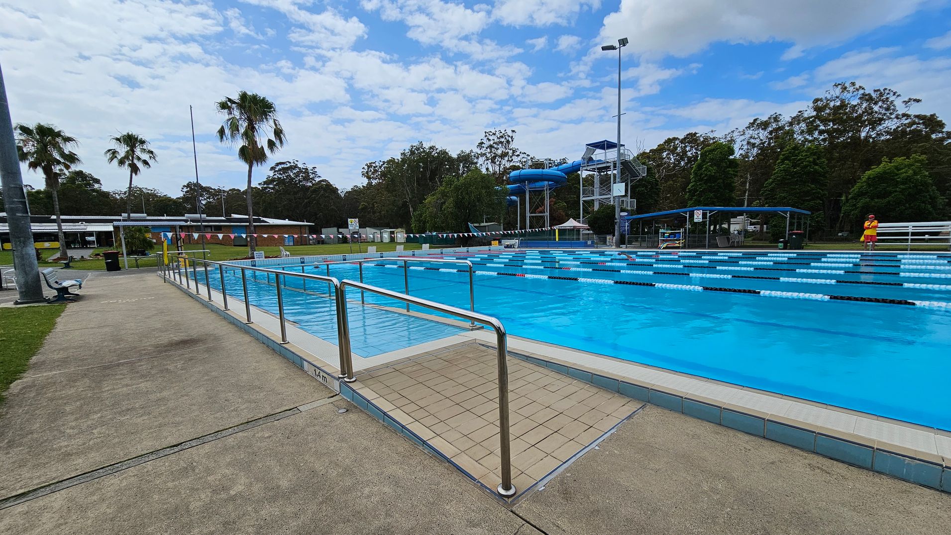 Outdoor swimming pool with marked lanes on the right, surrounded by a metal railing. A lifeguard in a red and yellow uniform stands near the pool. On the left, a paved walkway with benches and a grassy area. In the background, there are palm trees, a building with a low roof, and a blue water slide structure. Tall trees are visible beyond the pool area.