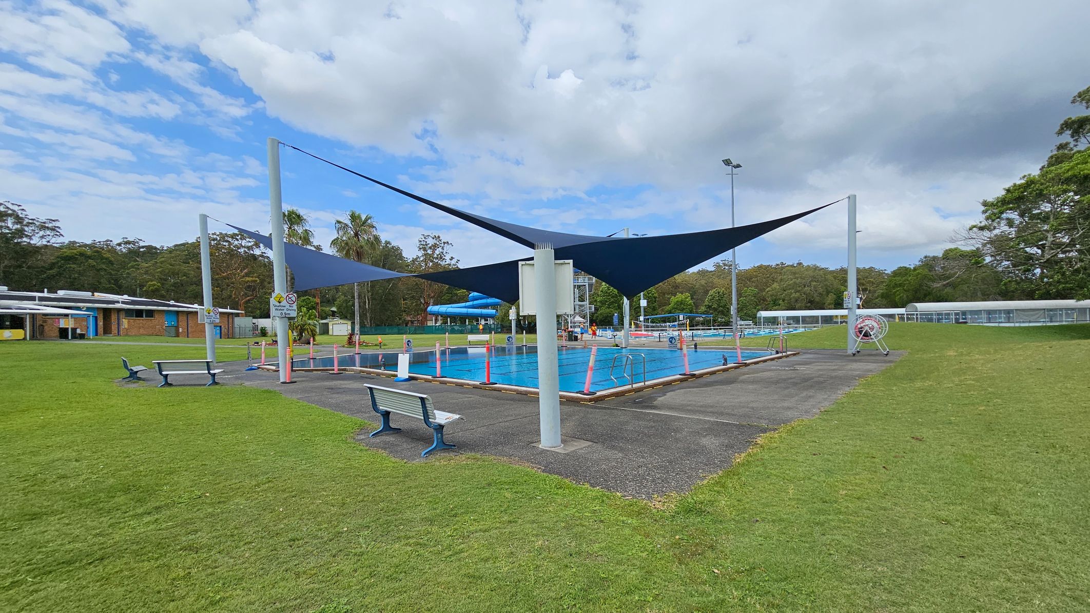 A swimming pool is in the centre, surrounded by a paved area with benches. Blue shade sails are suspended above the pool. On the left, there is a building with a blue and yellow facade. The right side features grassy areas and a covered walkway. Trees and greenery are visible in the background.