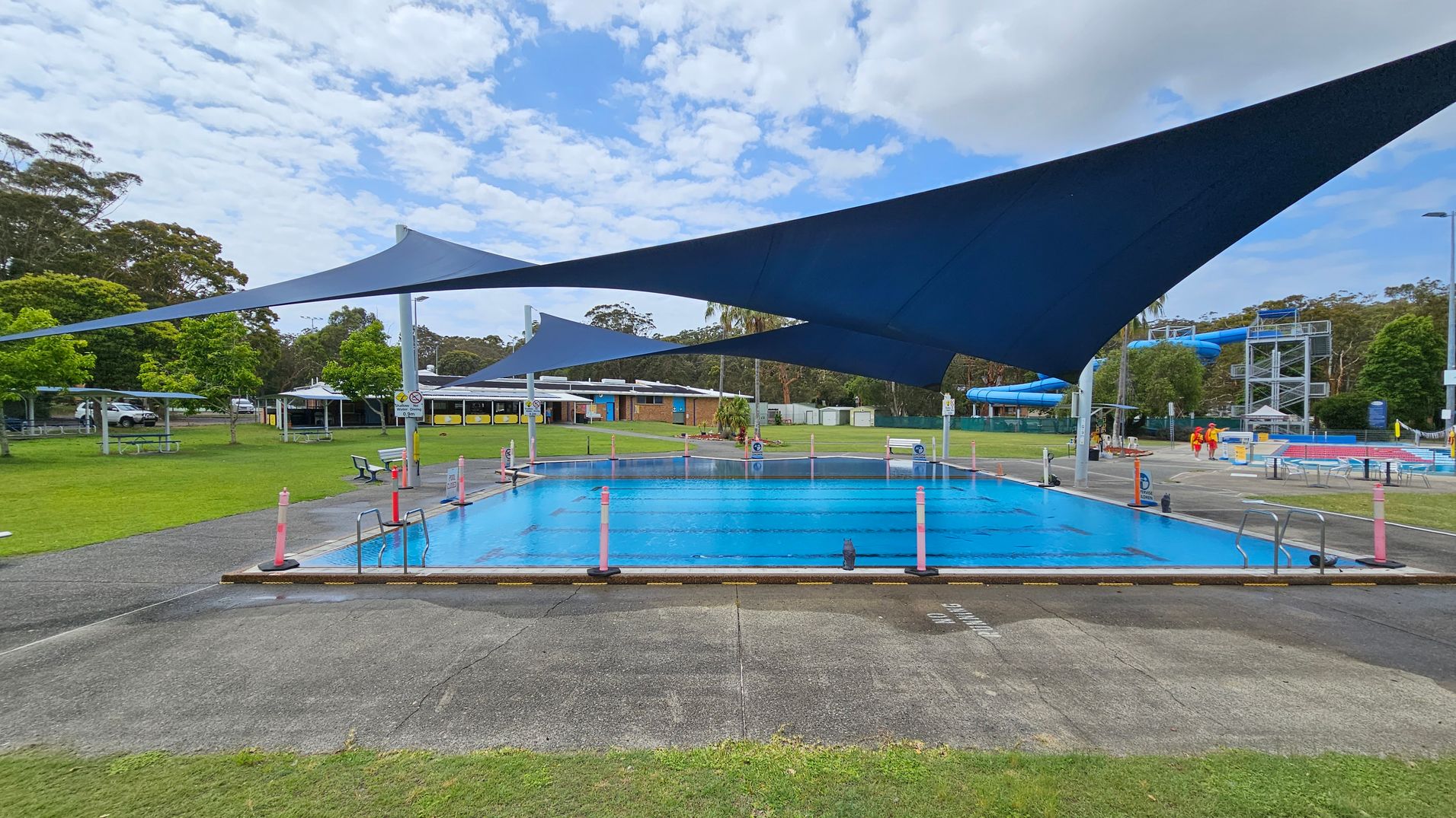 A swimming pool with blue water is in the centre, surrounded by a concrete path. Above, large blue shade sails provide cover. On the left, there are green lawns with picnic tables and trees. In the background, a building with a white roof is visible. On the right, two people in red and yellow uniforms walk near a blue water slide structure. The area is open and surrounded by greenery.