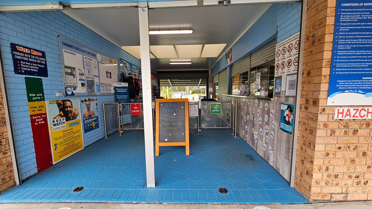 Entrance to a swimming facility with blue hexagonal floor tiles. On the left, a blue wall displays various safety and informational signs, including a "Wild Ride" notice. In the centre, a wooden board lists slide hours. On the right, a brick wall with a "Conditions of Entry" sign and a counter with closed shutters. Above, fluorescent lights illuminate the area. The entrance is flanked by metal barriers with "No Entry" and "Enter" signs.