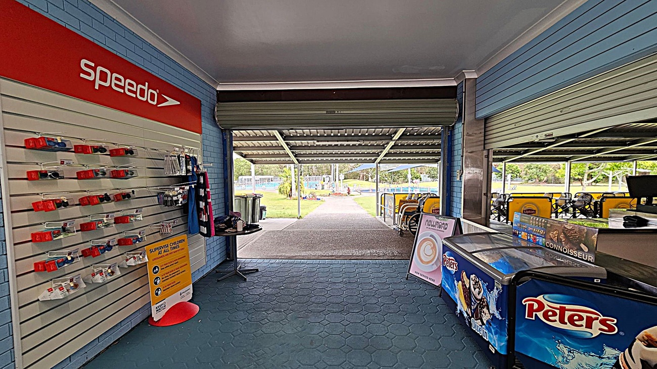 On the left, a display wall with Speedo-branded swimming accessories and a sign about supervising children. In the middle, an open area leads to an outdoor space with a path and greenery. On the right, a counter with a Peters ice cream freezer and a menu board. The floor is covered with hexagonal tiles, and the ceiling is plain.