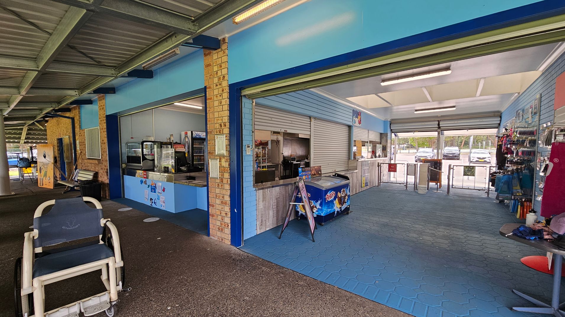 A covered outdoor area with a corrugated metal ceiling and a concrete floor. On the left, there's a wheelchair and a bench against a brick wall. In the centre, a kiosk with a blue counter displays a variety of items, including an ice cream freezer. The right side has a blue-tiled floor and shelves with merchandise. The background shows a car park visible through open roller doors.