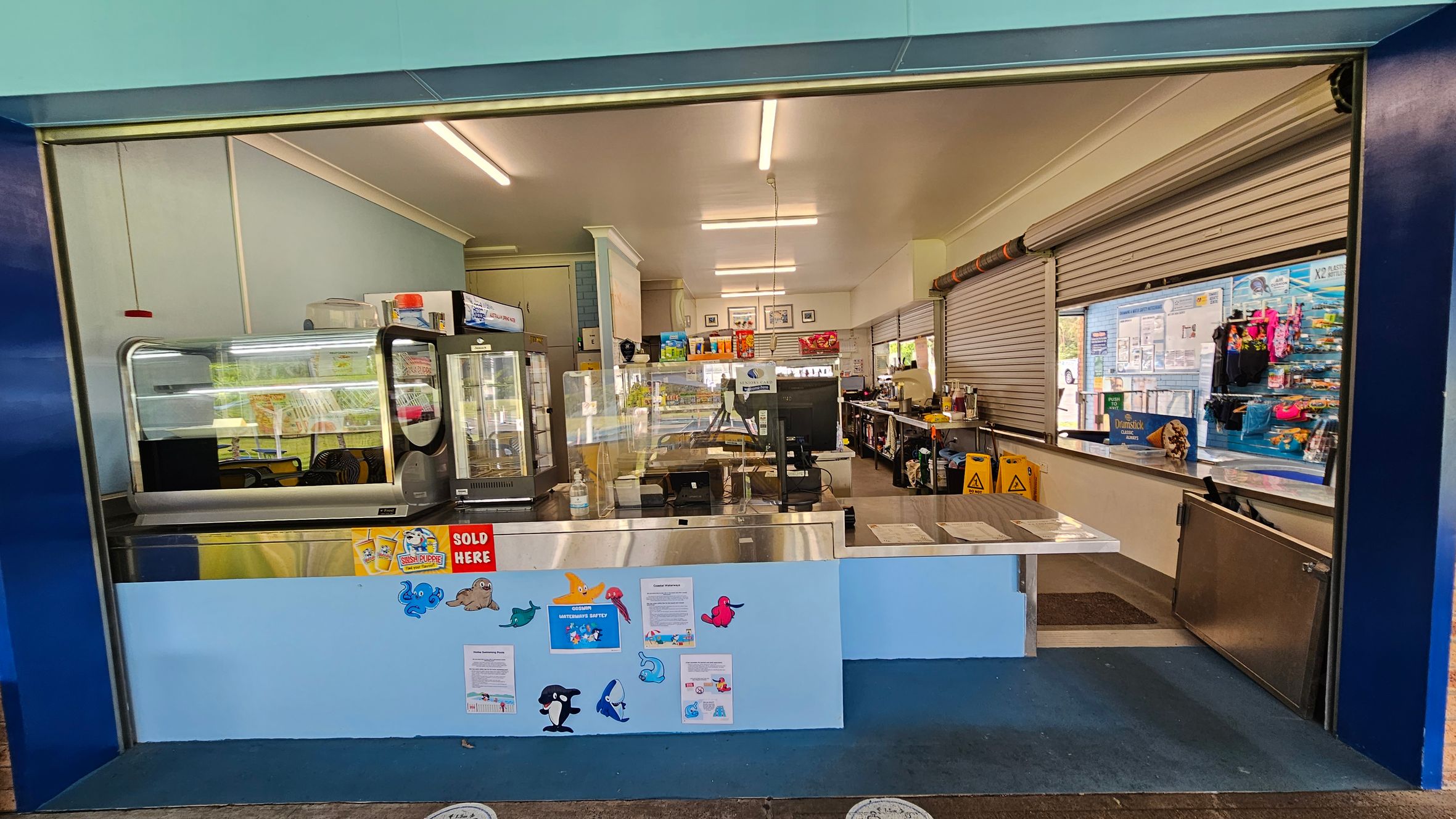 A canteen with a wide serving counter in the foreground, decorated with colourful sea creature cutouts and signs. Behind the counter, there are display fridges and shelves stocked with various items. The interior is well-lit with fluorescent lights, and the walls are light blue. On the right, there are closed roller shutters and a display of snacks and drinks. The floor is blue, and there are caution signs visible inside.