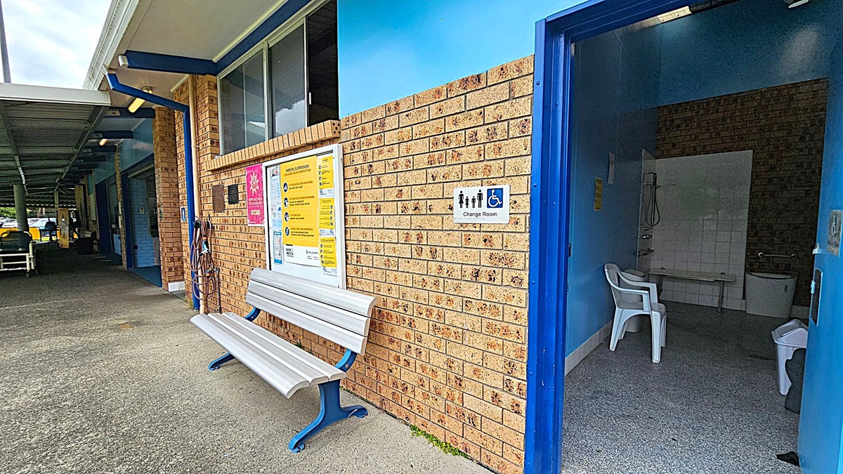 A brick building with a blue trim features a change room on the right, marked by a sign with accessibility symbols. Inside, there's a white plastic chair and a tiled area with a shower. On the left, a white bench with blue legs is positioned against the wall, which displays various informational posters. The floor is concrete, and a covered walkway extends into the background.