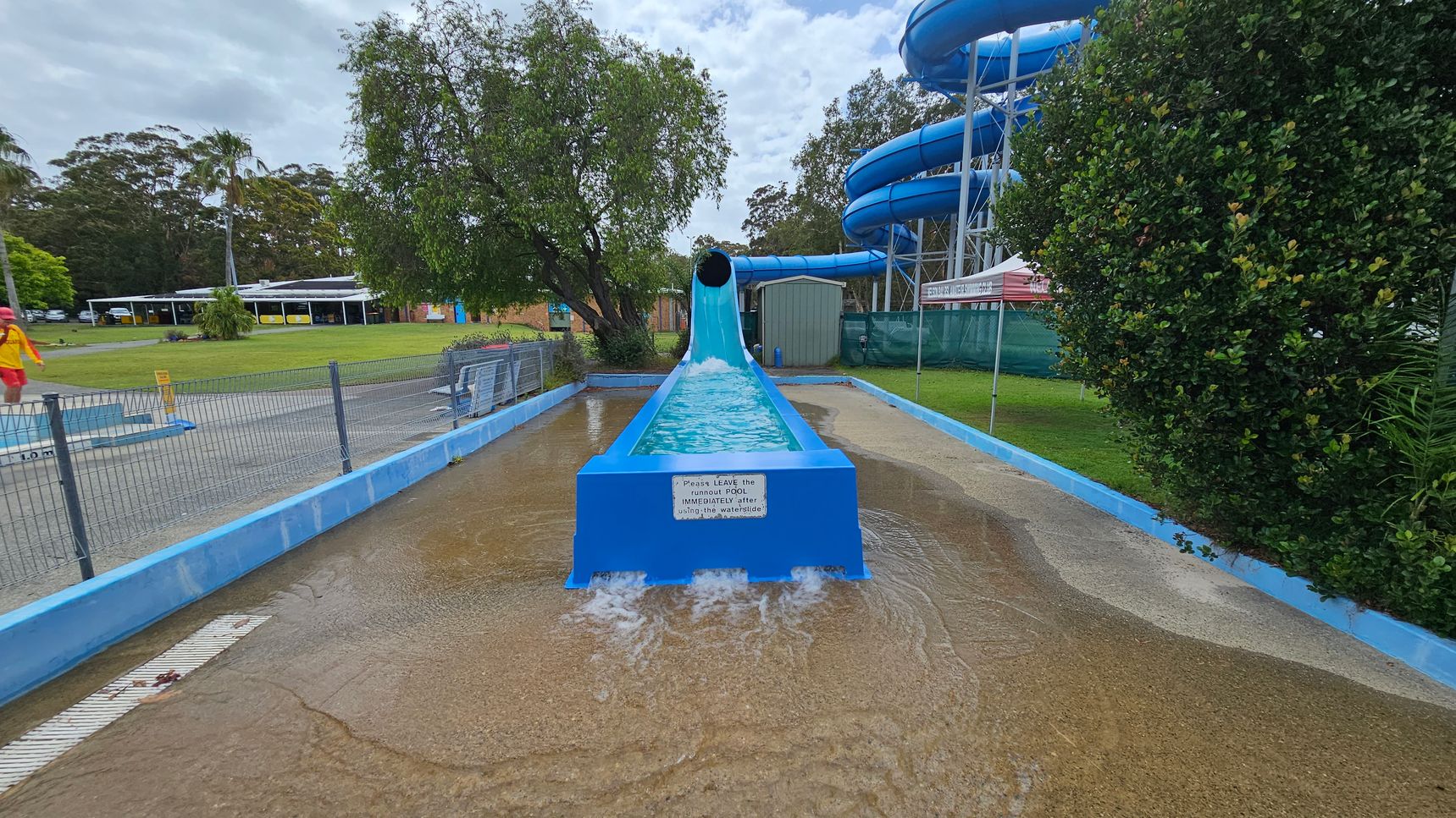 A blue water slide with a tunnel at the top ends in a shallow pool. Water flows out of the slide into the pool. On the left, a lifeguard in a red and yellow uniform walks near a fenced area. In the background, there are trees and a building with a white roof. On the right, a large bush and a blue spiral slide structure are visible. The ground is wet, and there is a sign on the slide's end.