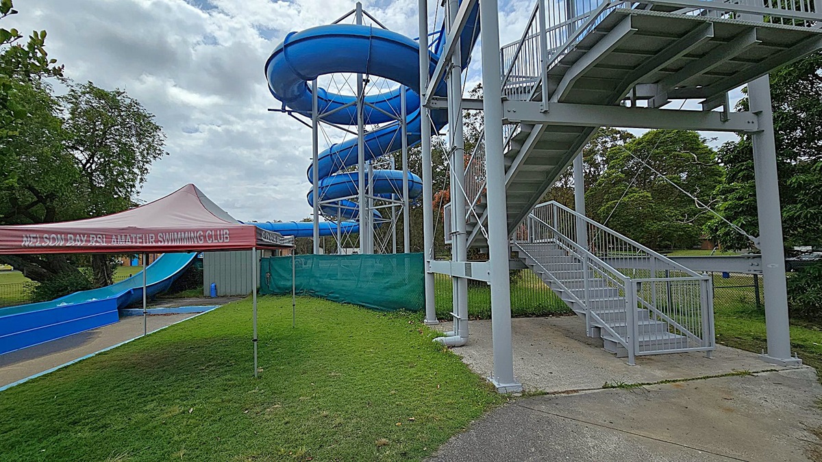 On the left, a blue water slide spirals down from a tall metal structure. Below it, a tent with "Nelson Bay RSL Amateur Swimming Club" written on it is set up on a grassy area. On the right, a metal staircase leads up to the slide, with a concrete path at its base. Trees and greenery are visible in the background, providing a natural setting around the water slide and tent.