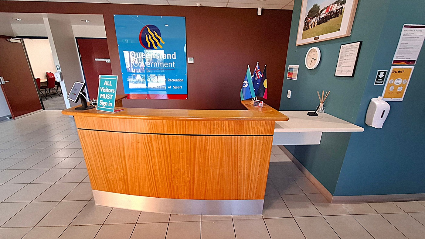 A reception area with a wooden desk in the centre. On the desk, there's a sign reading "ALL Visitors MUST Sign in" and a tablet. Behind the desk, a blue sign with "Queensland Government" is mounted on a brown wall. To the right, three small flags are displayed. The wall also features a clock, framed picture, and various notices. The floor is tiled, and a hand sanitiser dispenser is mounted on the wall. An open door to the left reveals a room with chairs.