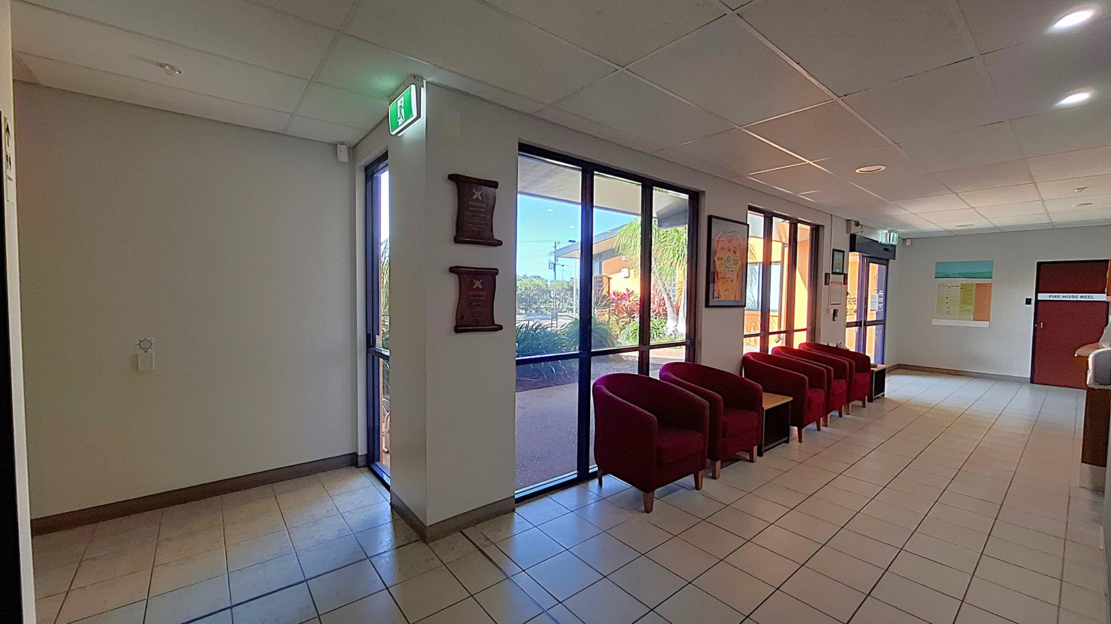 A waiting area with a tiled floor and a grid-patterned ceiling. On the left, there are large glass doors with an illuminated exit sign above. Two plaques are mounted on the wall beside the doors. In the middle, a row of red upholstered chairs is aligned against the wall, with small tables between them. On the right, a door marked "Fire Hose Reel" is visible, along with a bulletin board displaying various notices.