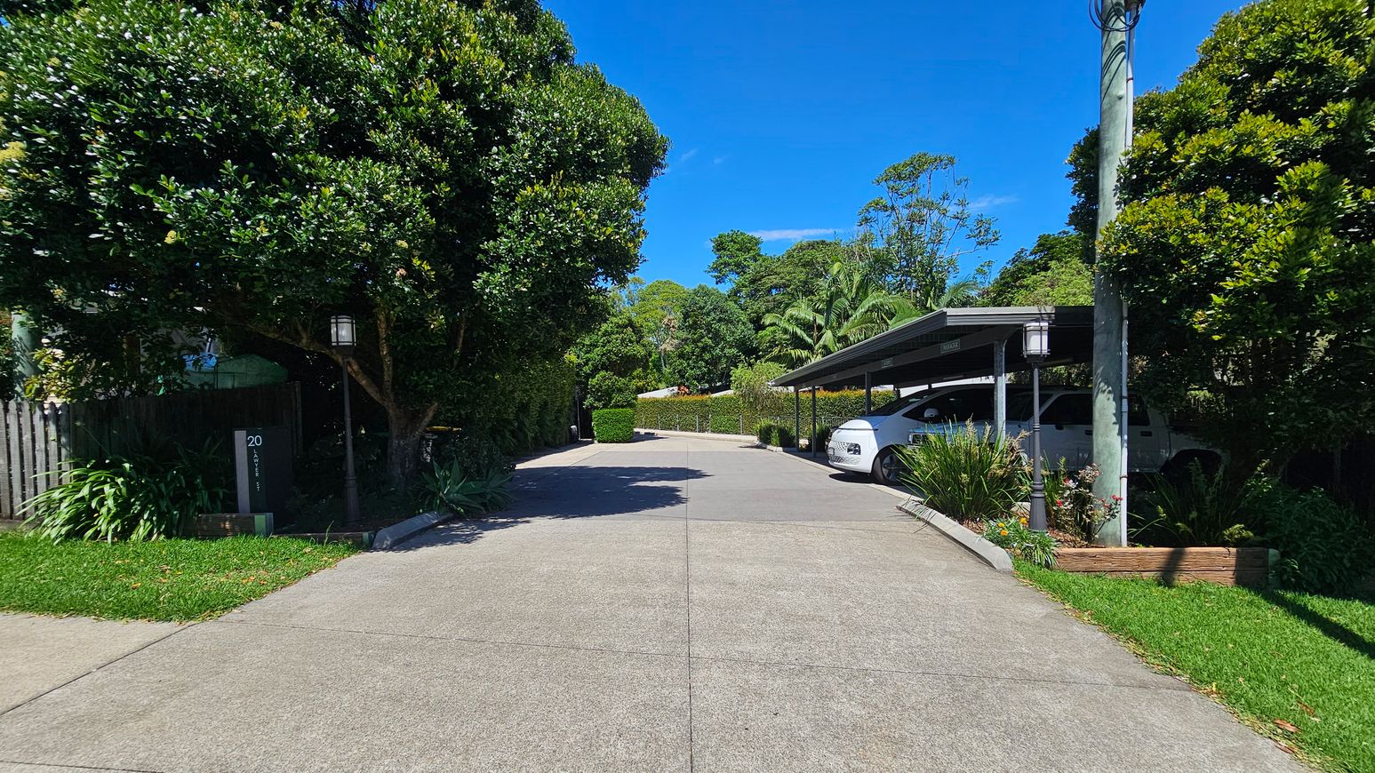 A driveway with a concrete surface leads to a covered parking area on the right, where a white car is parked. On the left, a large tree and a wooden fence are visible, with a house number sign marked "20". The area is surrounded by lush greenery, including bushes and trees. A lamp post stands near the parking area, and a power pole is visible. The scene is well-lit, suggesting a sunny day.
