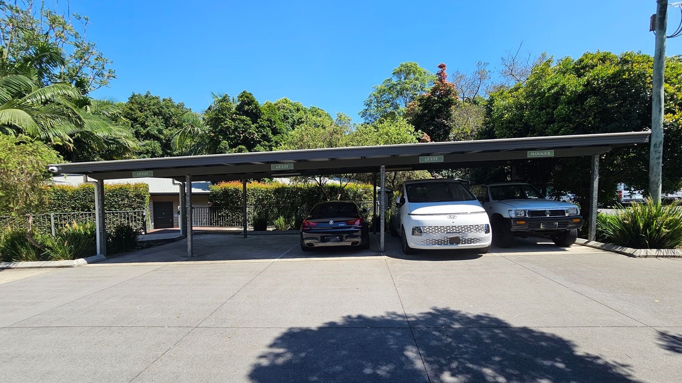 A covered parking area with three vehicles parked side by side. On the left, a black car is parked under a sign marked "GUEST". In the middle, a white van is parked under another "GUEST" sign. On the right, a grey vehicle is parked under a "MANAGER" sign. Behind the parking area, there is a hedge and a building with a metal roof. Lush green trees are visible in the background. The ground is paved with concrete, and there are shadows from the trees.