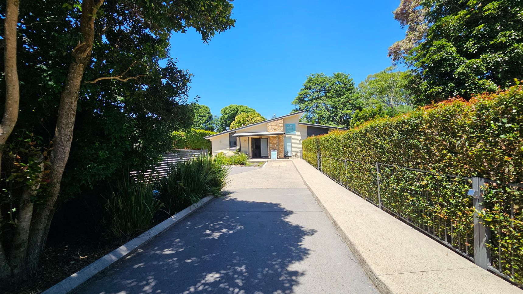 A modern, single-storey building with a slanted roof is at the end of a paved driveway. On the left, there are tall trees and bushes, while on the right, a neatly trimmed hedge lines the path. The building features a mix of stone and light-coloured walls, with a sign above the entrance. Lush greenery surrounds the area, creating a serene atmosphere.